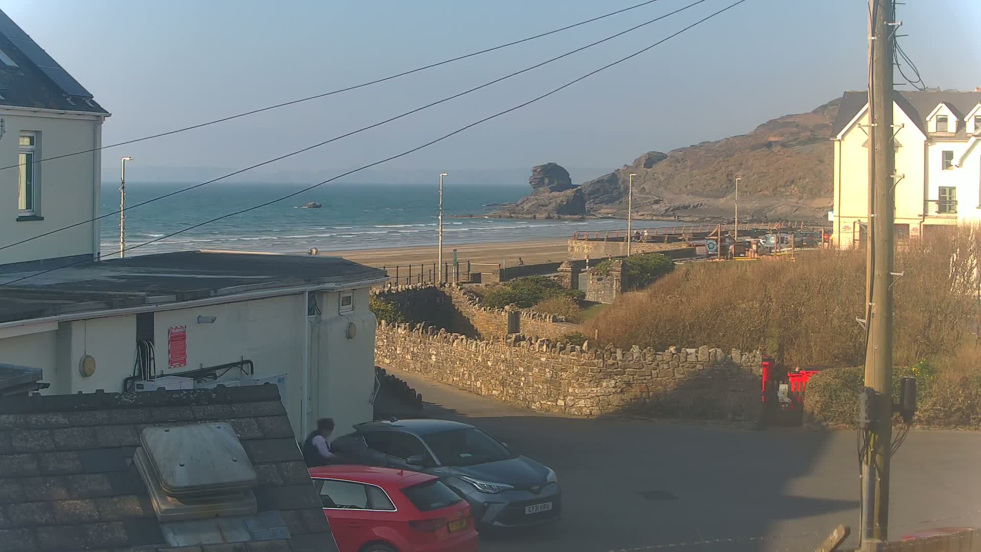A sunny coastal scene shows a sandy beach with gentle waves, a rocky headland, buildings, and parked cars; a stone wall separates the street from the beach.