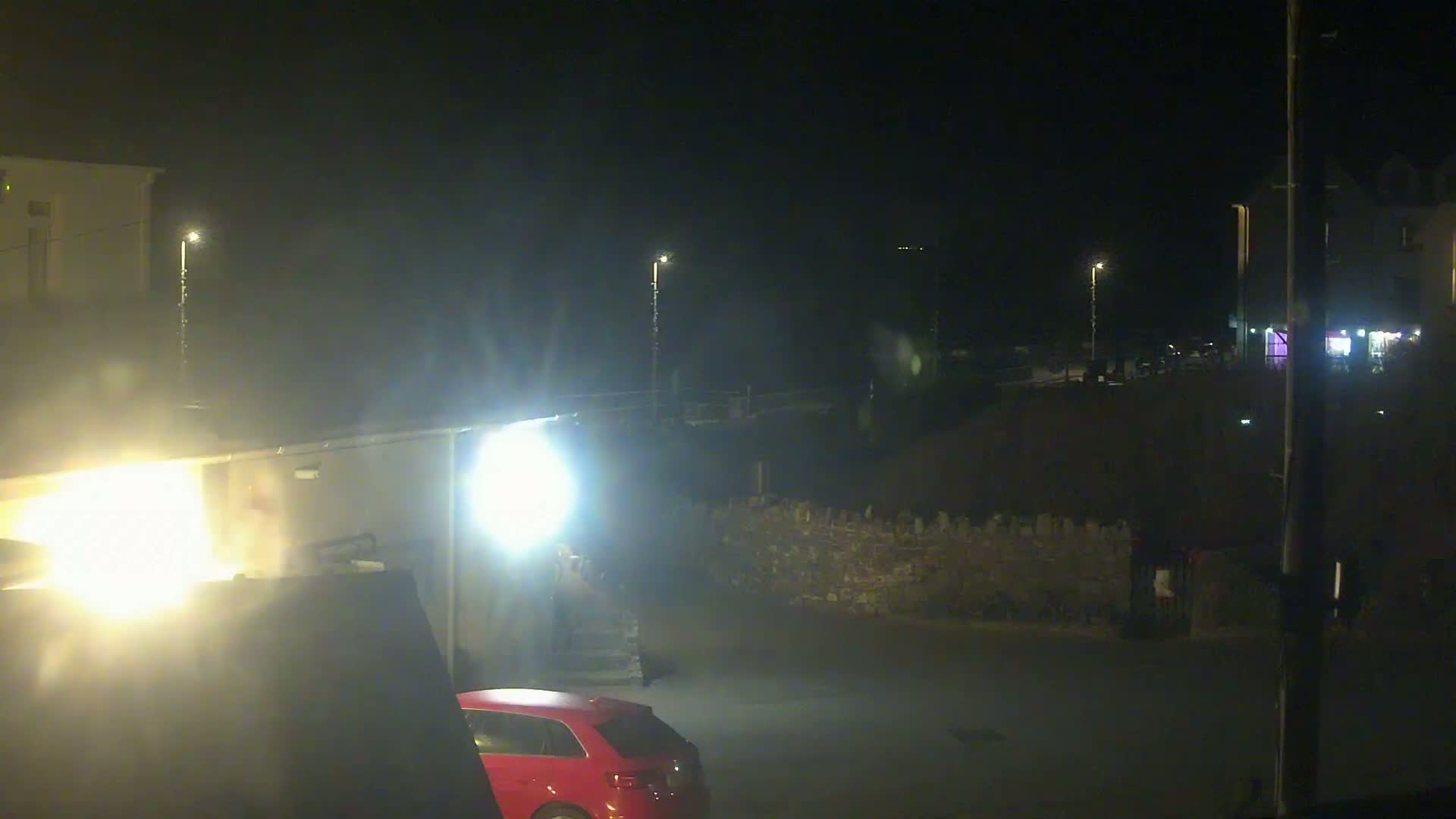 A nighttime, dimly lit street scene shows a red car parked near a building, with a stone wall and streetlights visible in the background.