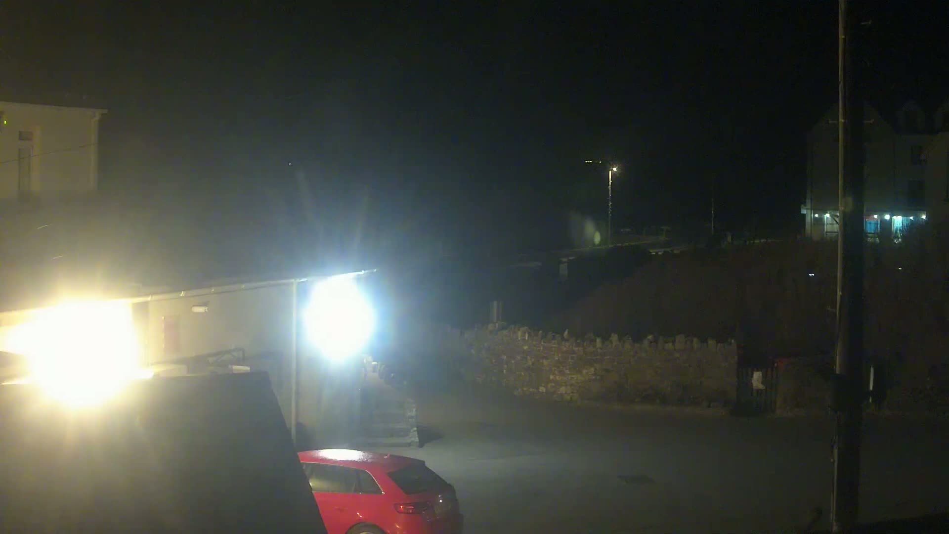 A red car is parked in a dark, nighttime outdoor scene illuminated by bright lights, with buildings and a low stone wall visible in the background.