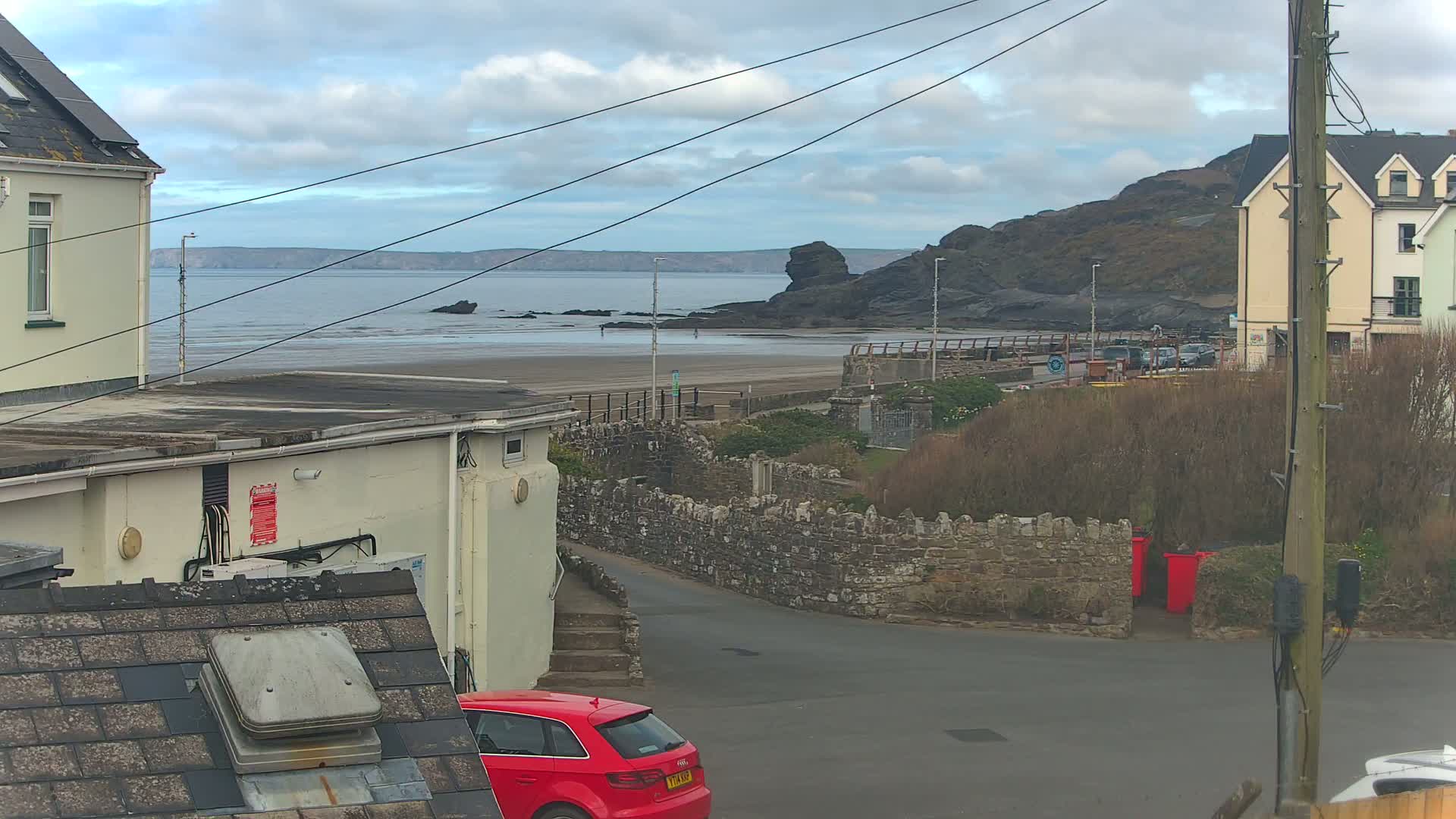 A view from a residential area shows a sandy beach with dark rocks and calm water under a cloudy sky, with a red car parked in the foreground.