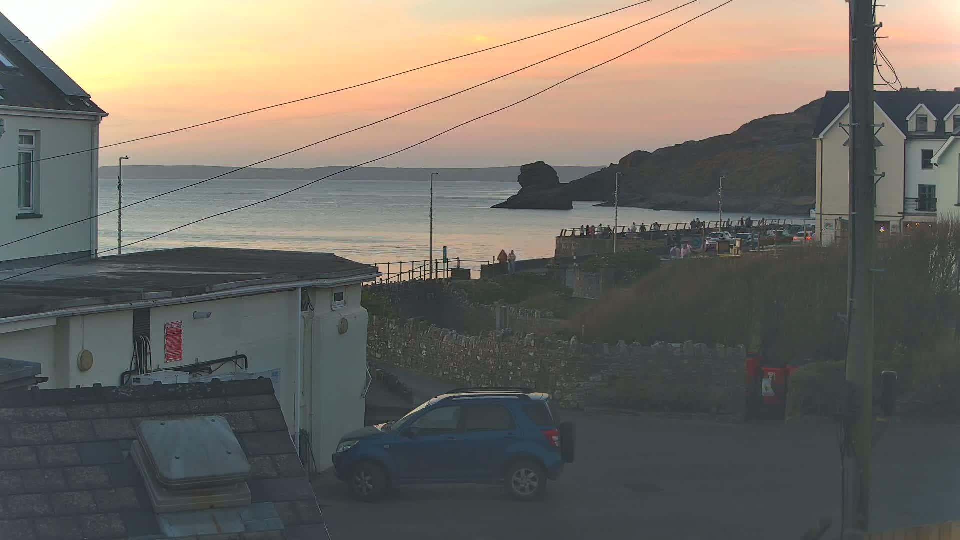 A blue SUV is parked on a street near a coastal town at sunset, with people gathered on a walkway overlooking the calm ocean and rocky cliffs.