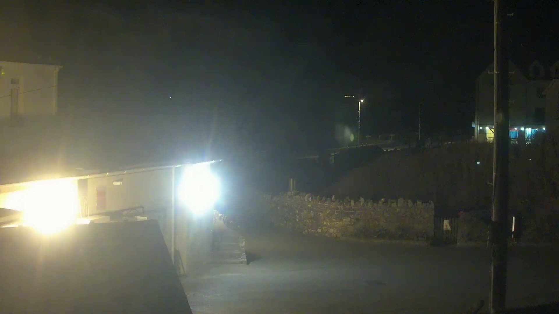 A nighttime scene shows a dimly lit street with buildings and a low stone wall, under hazy conditions.