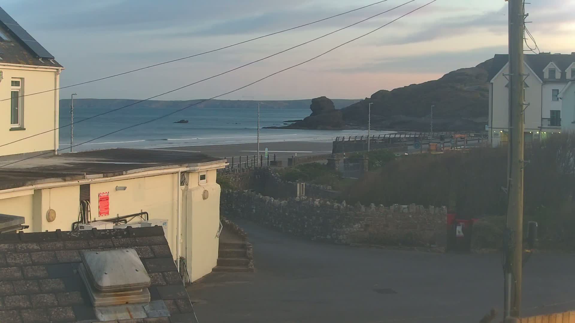 A view from a building shows a calm ocean beach under a partly cloudy sky at dusk, with buildings and a stone wall in the foreground.