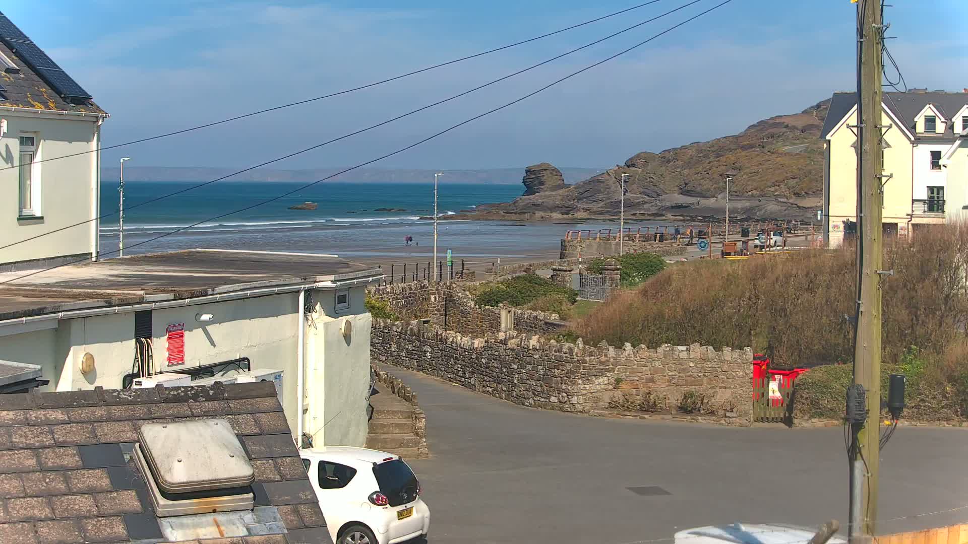 A sunny coastal scene shows a sandy beach with small waves, a rocky outcrop, buildings, and a white car parked near a stone wall.