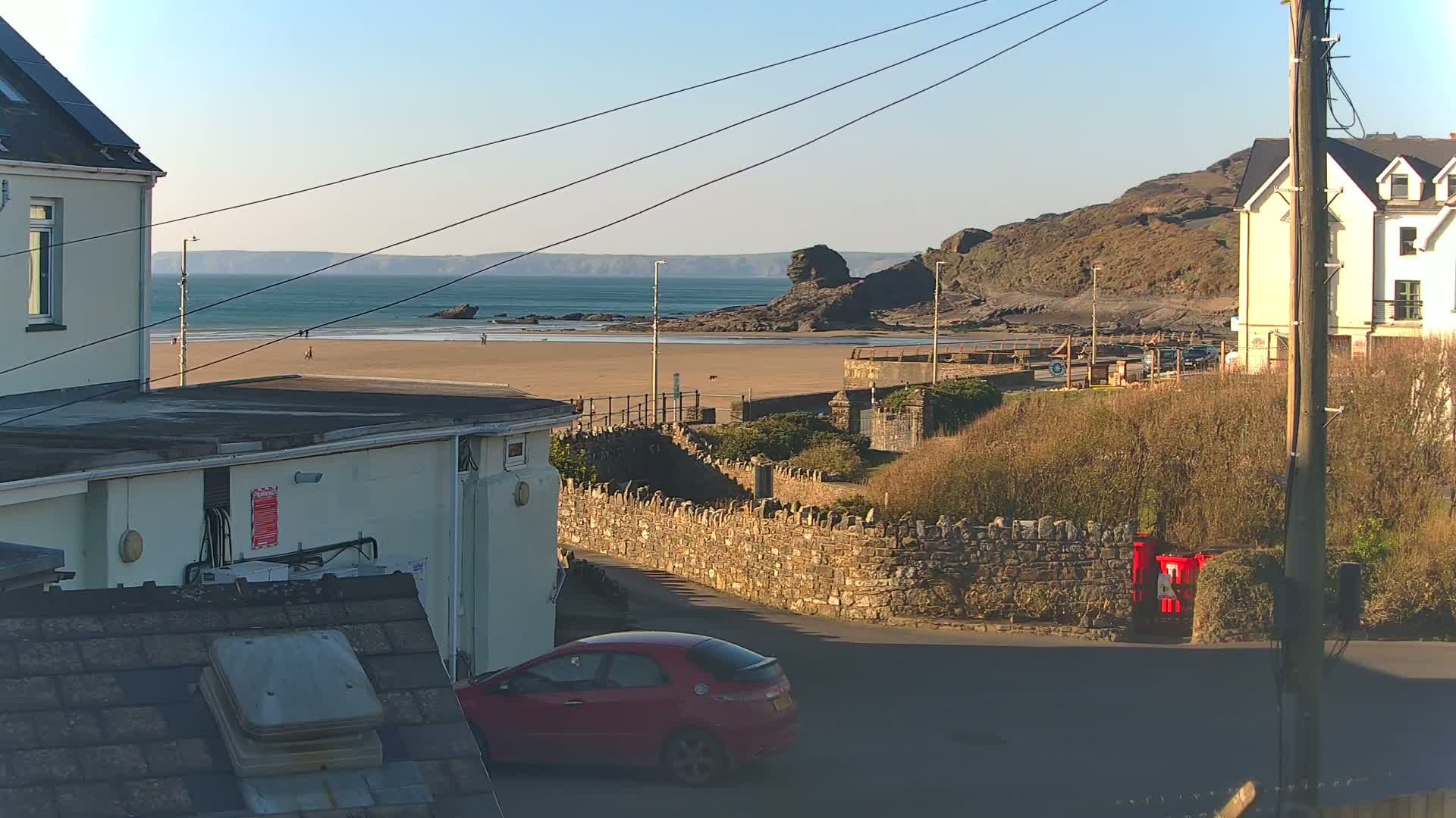 A red car is parked on a street overlooking a sandy beach, calm ocean, and rocky cliffs under a clear, sunny sky.