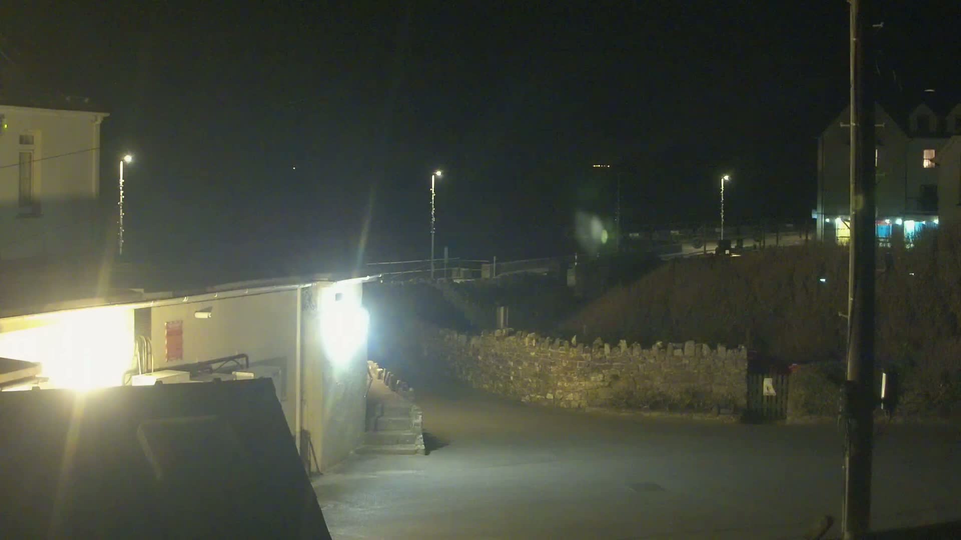 A nighttime view of a dimly lit street scene with buildings, a low stone wall, and bright security lights.