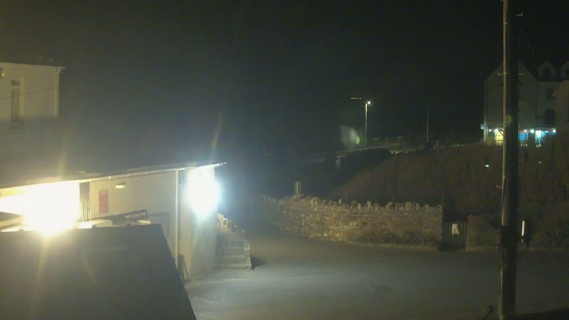 A nighttime, dimly lit scene shows a paved area between buildings and a low stone wall, with a few people visible near a building.