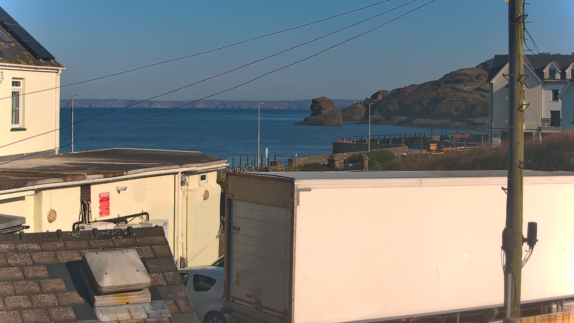 A large white delivery truck is parked in front of a coastal scene under a clear blue sky, with a calm ocean and rocky coastline in the background.