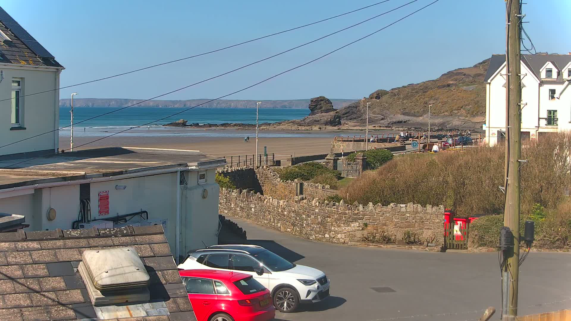 A sunny beach scene is viewed from a residential area, showing two parked cars, buildings, and a stone wall overlooking a sandy beach with a calm ocean in the background.
