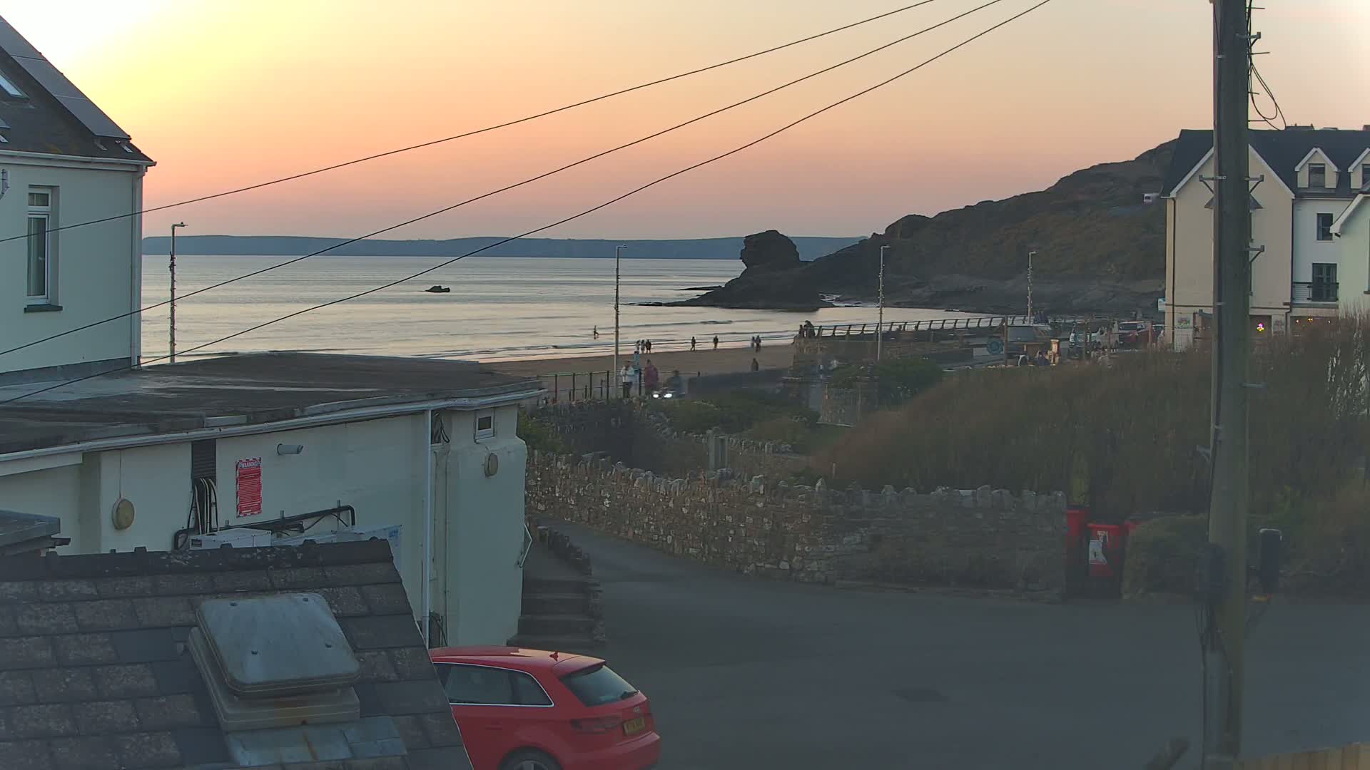 A sunset over a calm ocean beach, viewed from a residential area with houses and a red car parked nearby.