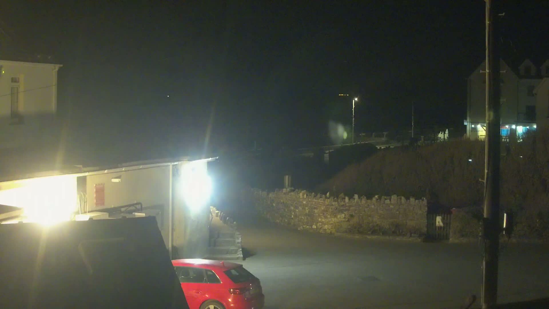 A red car is parked in a dimly lit area at night near buildings and a low stone wall.