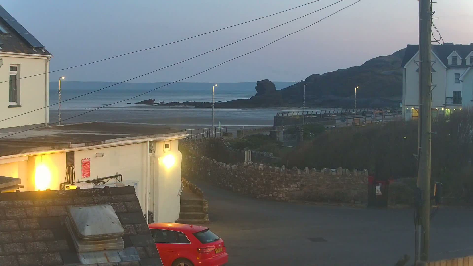 A red car is parked near a building overlooking a calm, twilight sea with a rocky shoreline and a low-lying hill in the background.