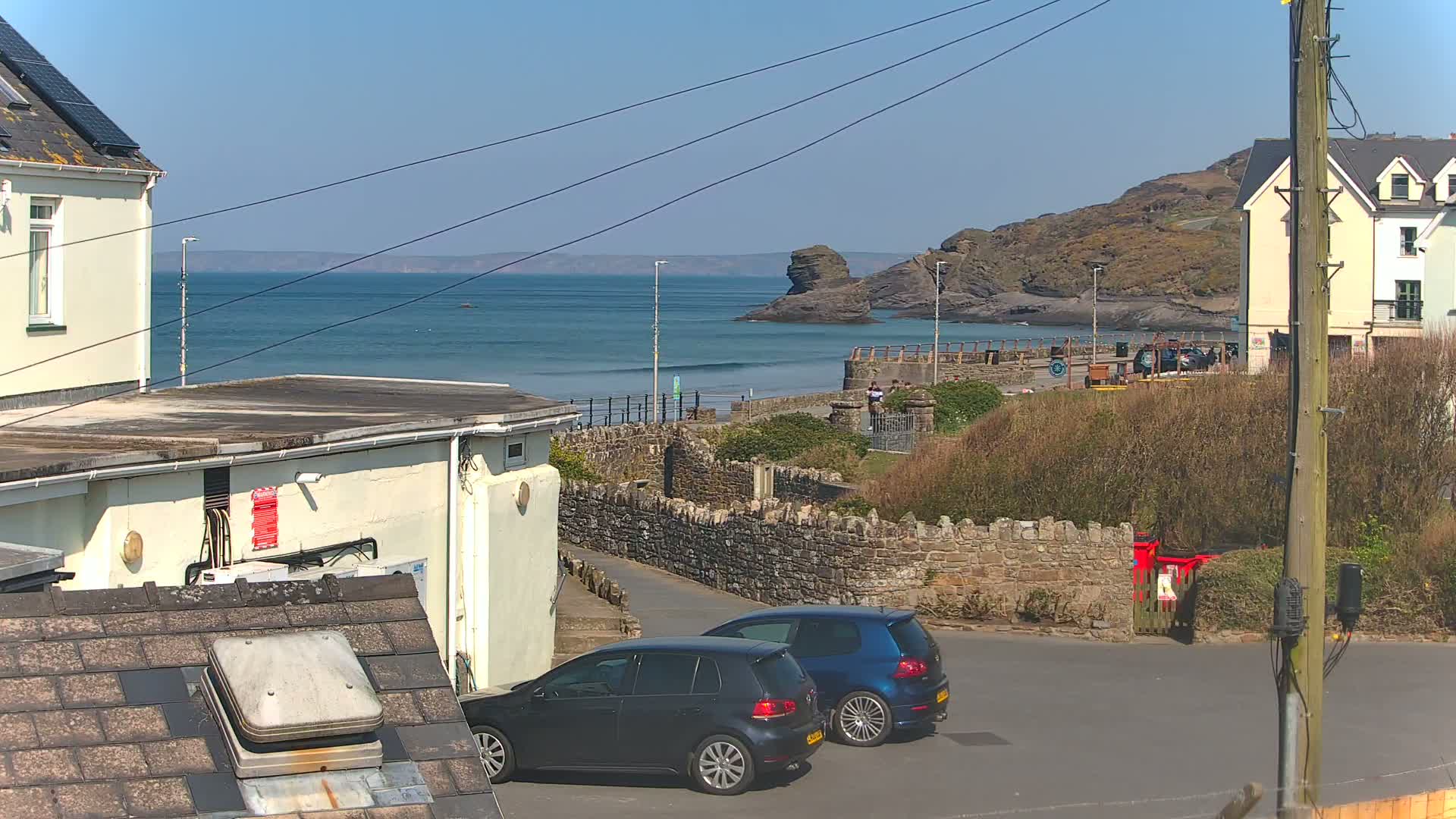 Two dark-colored cars are parked in a small seaside town parking lot on a sunny day, overlooking a calm ocean with a rocky coastline.