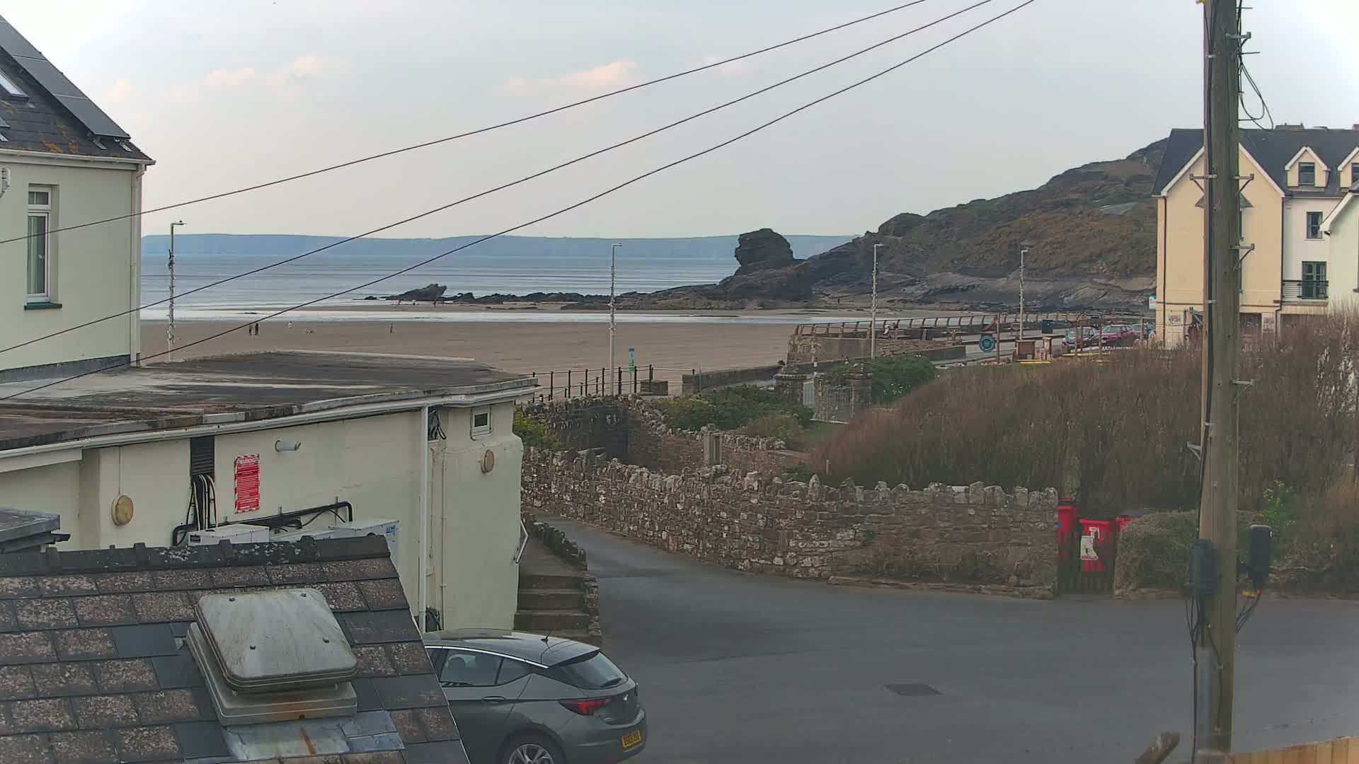 A view from a residential area shows a sandy beach, calm ocean, and rocky outcropping under an overcast sky; a car is parked on a street below.