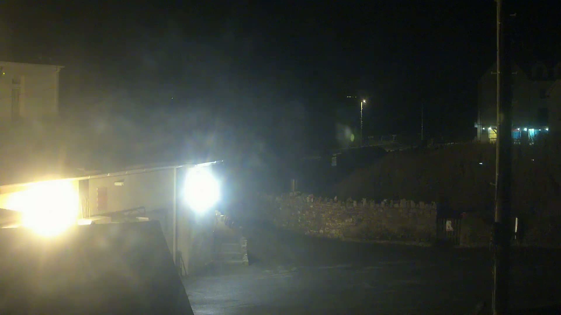 A foggy, dark night scene shows a dimly lit street with buildings, a stone wall, and bright lights illuminating a portion of the foreground.