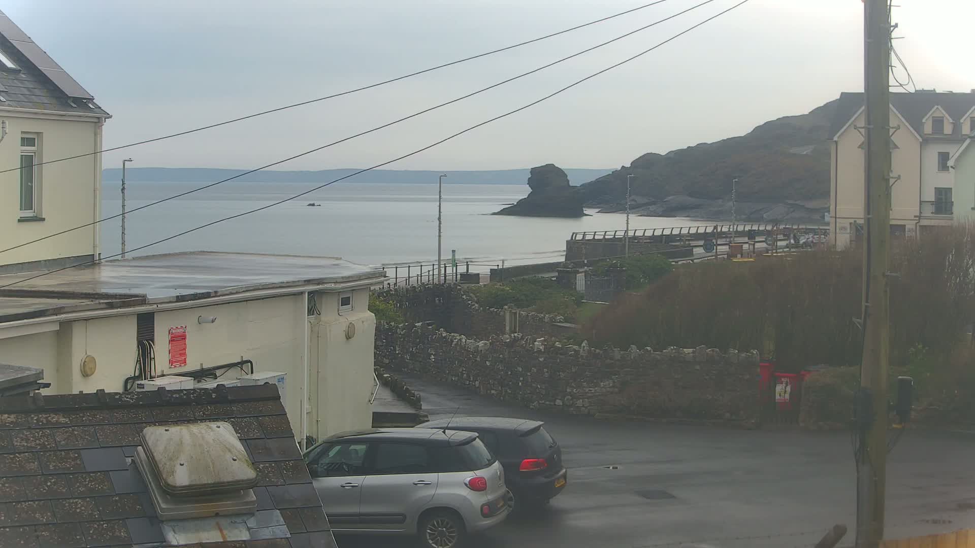 A wet, overcast day reveals a coastal view of a calm sea, a rocky outcrop, a beach, buildings, and two parked cars from an elevated vantage point.