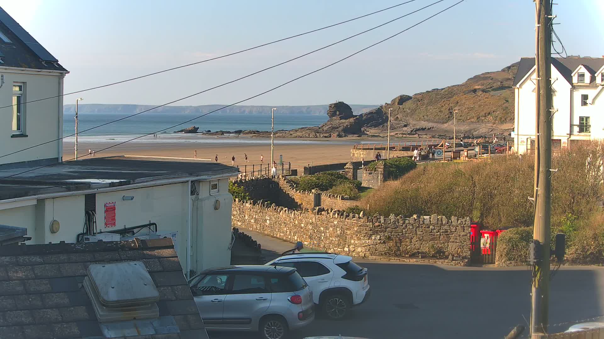 A sandy beach with a few people on it is visible from a seaside town under a clear, sunny sky; two cars are parked on a street leading toward the beach.