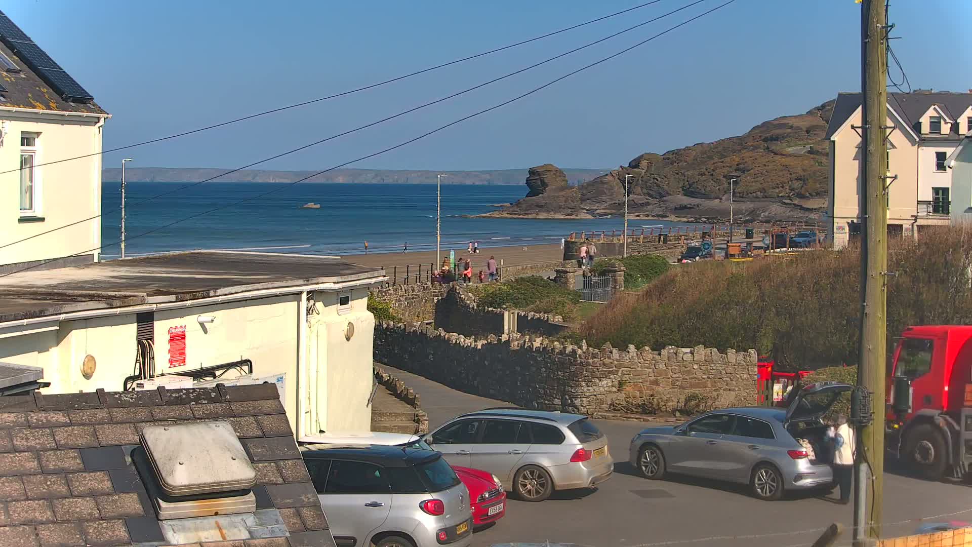 A sunny coastal scene shows a sandy beach with a few people, several parked cars, and buildings near a stone wall, all overlooking a calm sea.
