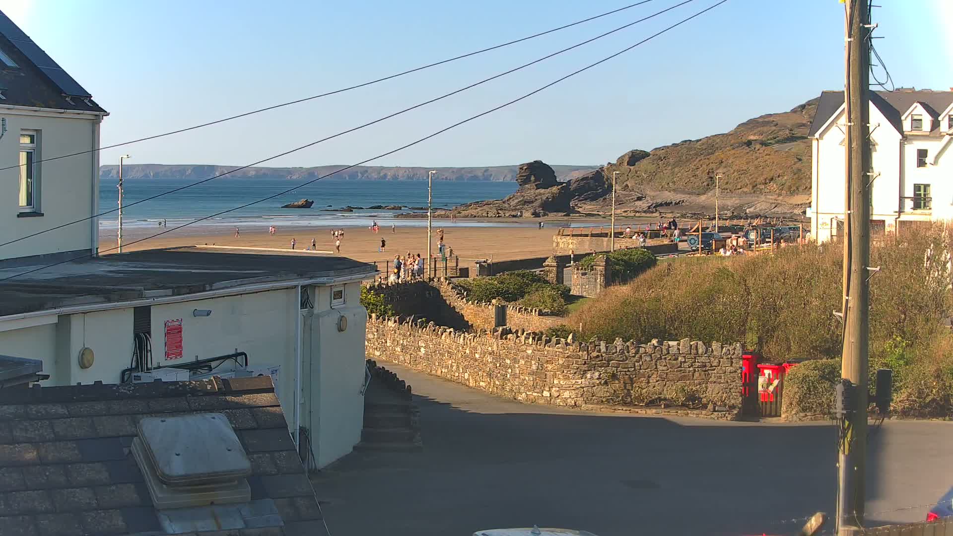 A sunny view of a sandy beach with people on it, backed by a stone wall and buildings, overlooking a calm ocean.