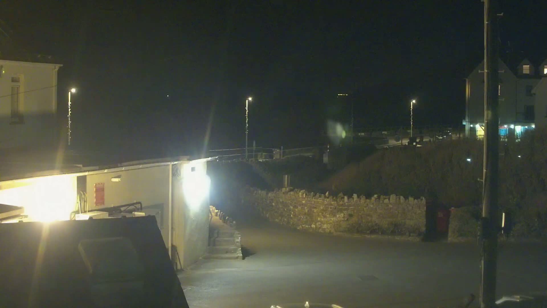 A nighttime view of a dimly lit street scene with buildings, a low stone wall, and streetlights.