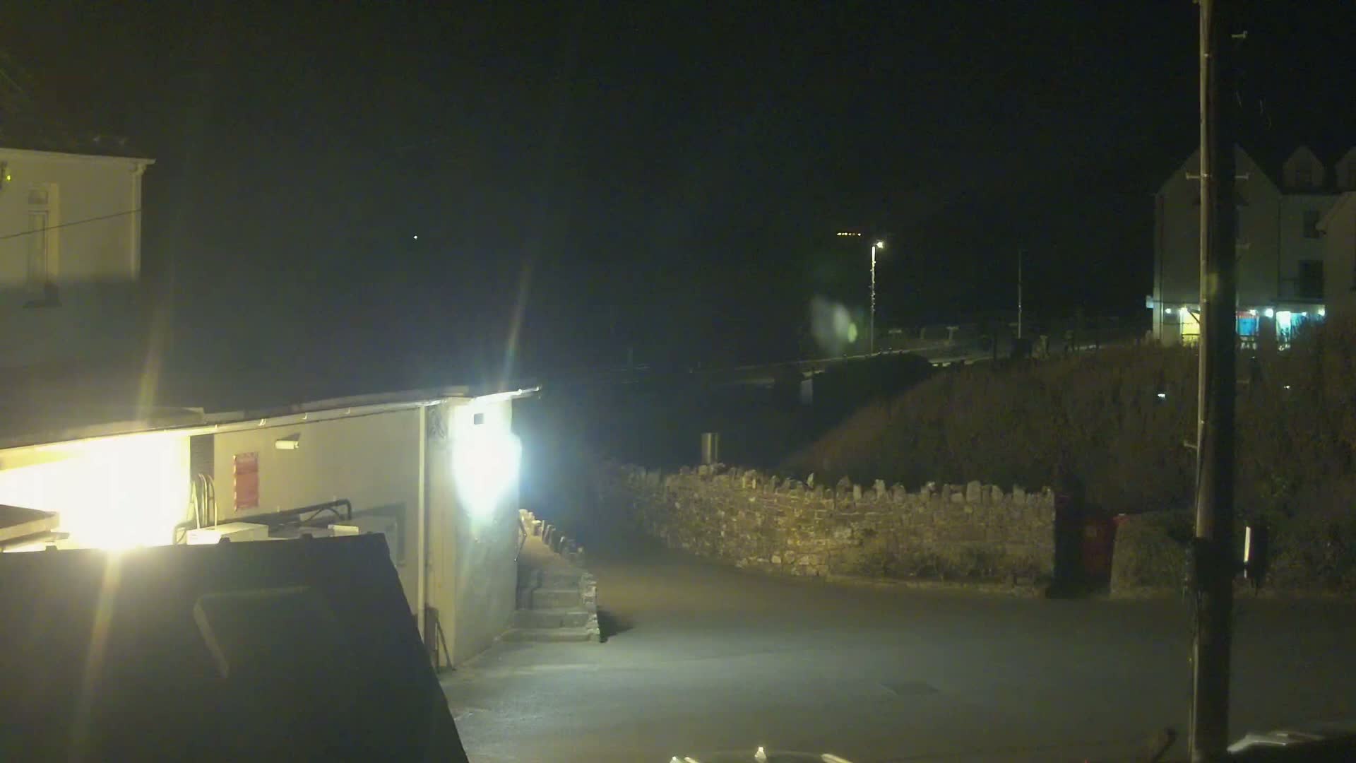 A nighttime view of a dimly lit street scene with buildings, a low stone wall, and a small parking area.
