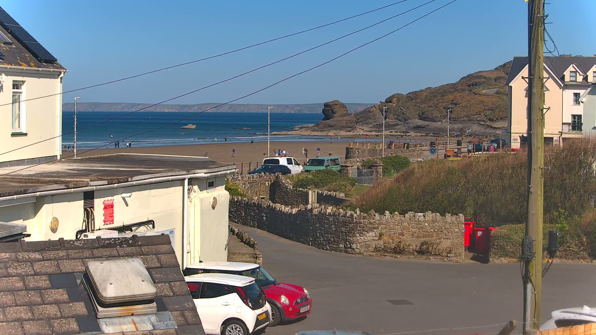A sandy beach with a few people on it is visible beyond a seaside town on a sunny day, with cars parked near stone walls and buildings.