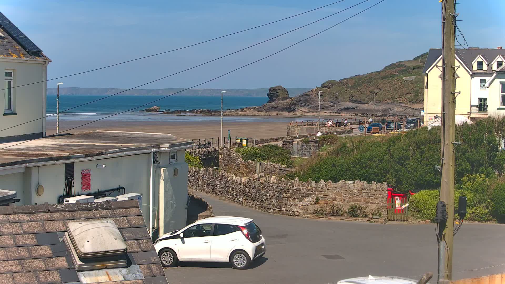 A white car is parked near a stone wall overlooking a sandy beach with a calm ocean under a clear, sunny sky.