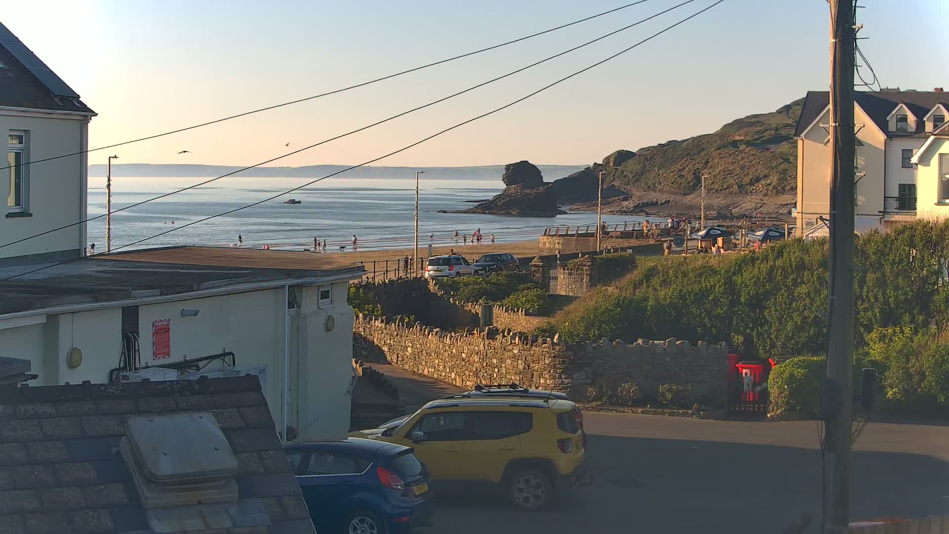 A seaside scene at sunset shows a sandy beach with people, a rocky outcropping in the water, houses, and cars parked on a street overlooking the ocean.