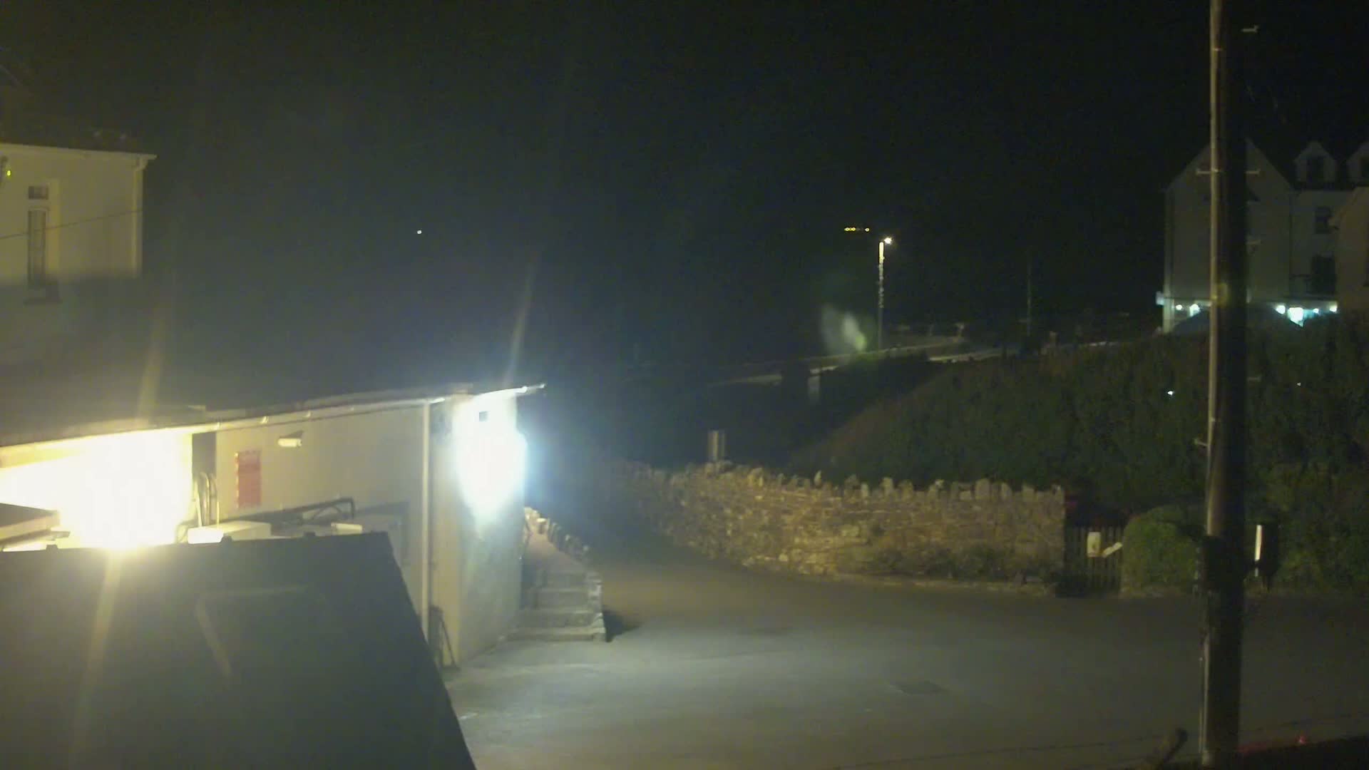 At night, a brightly lit area shows a building, a low stone wall, and a street, all under a dark sky.