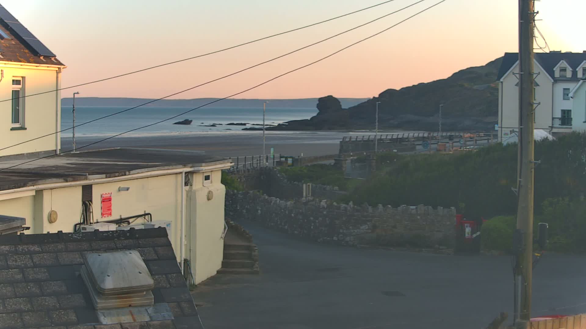 A coastal scene at sunset shows a sandy beach, calm ocean, and buildings nestled against a rocky hillside.