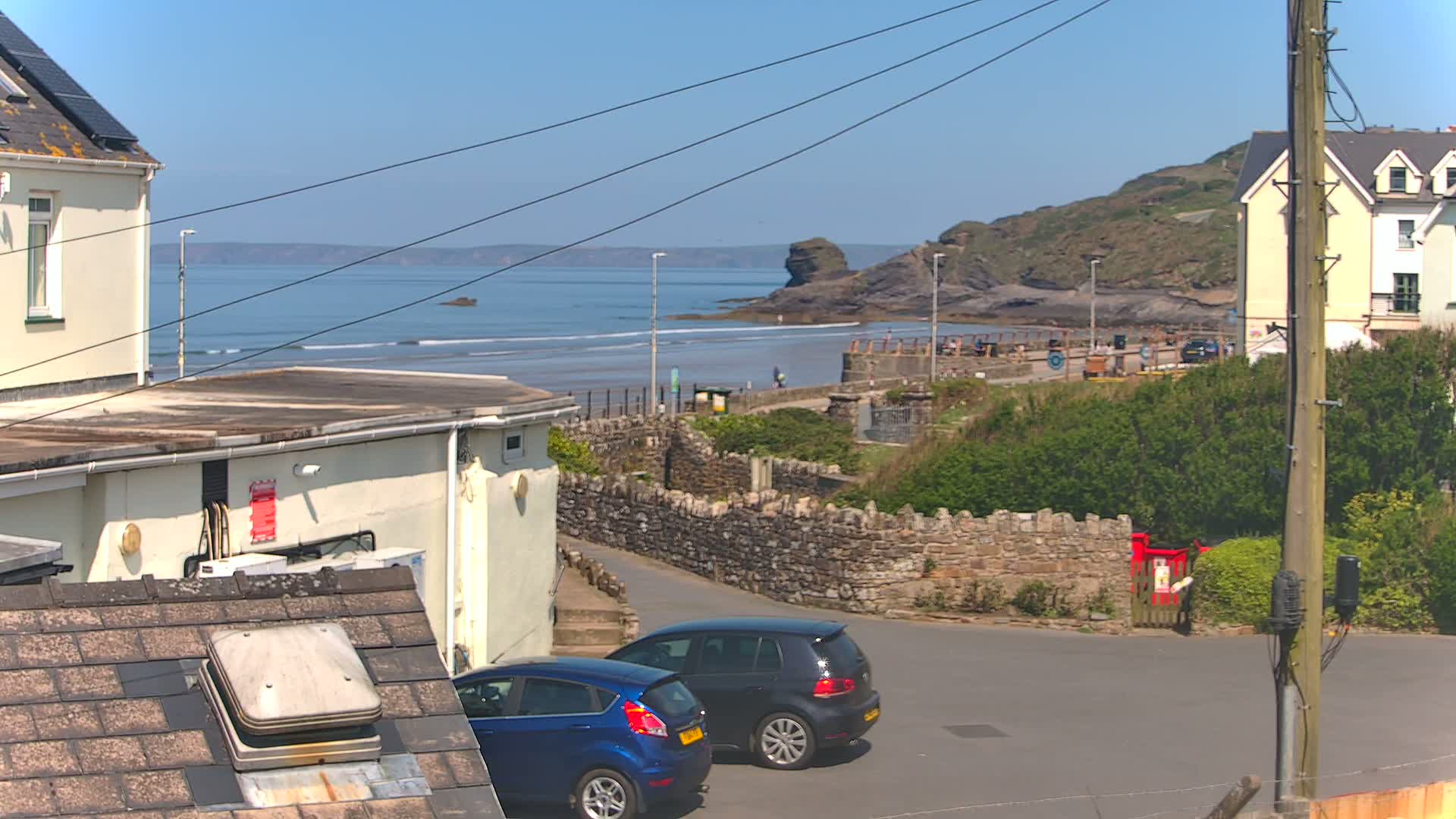 Two cars are parked near a stone wall overlooking a beach with gentle waves under a clear, sunny sky.
