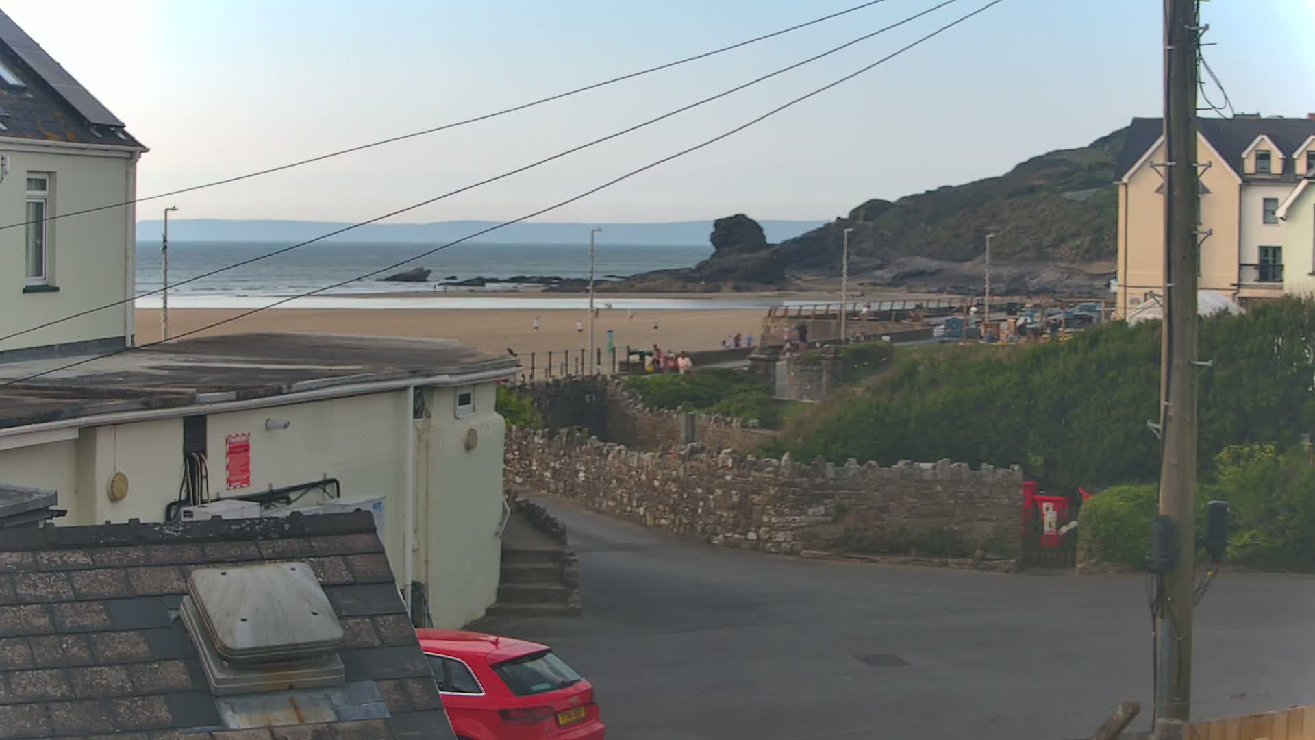 A view of a sandy beach with people on it, under a mostly clear sky, is seen from between buildings, with a red car parked nearby.