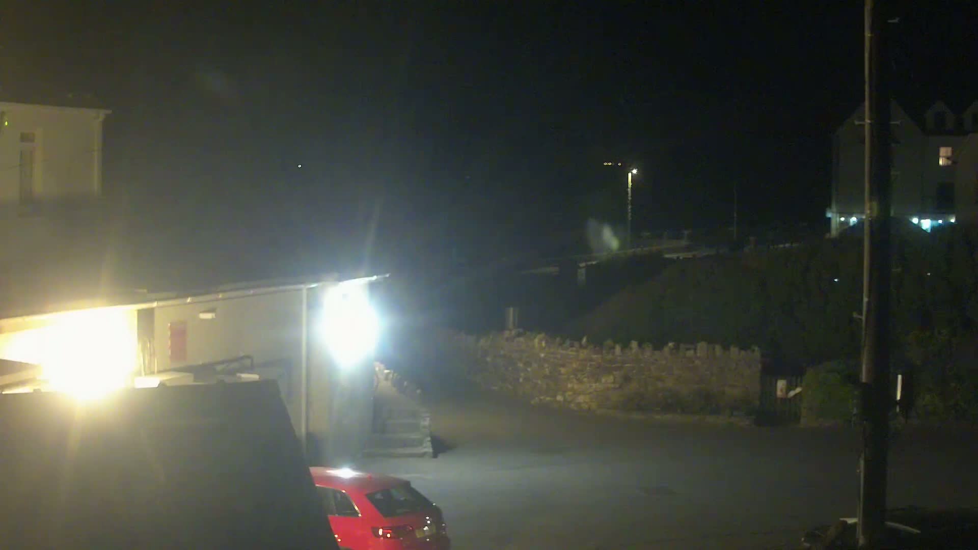 A red car is parked in a dimly lit area at night, near buildings and a low stone wall.