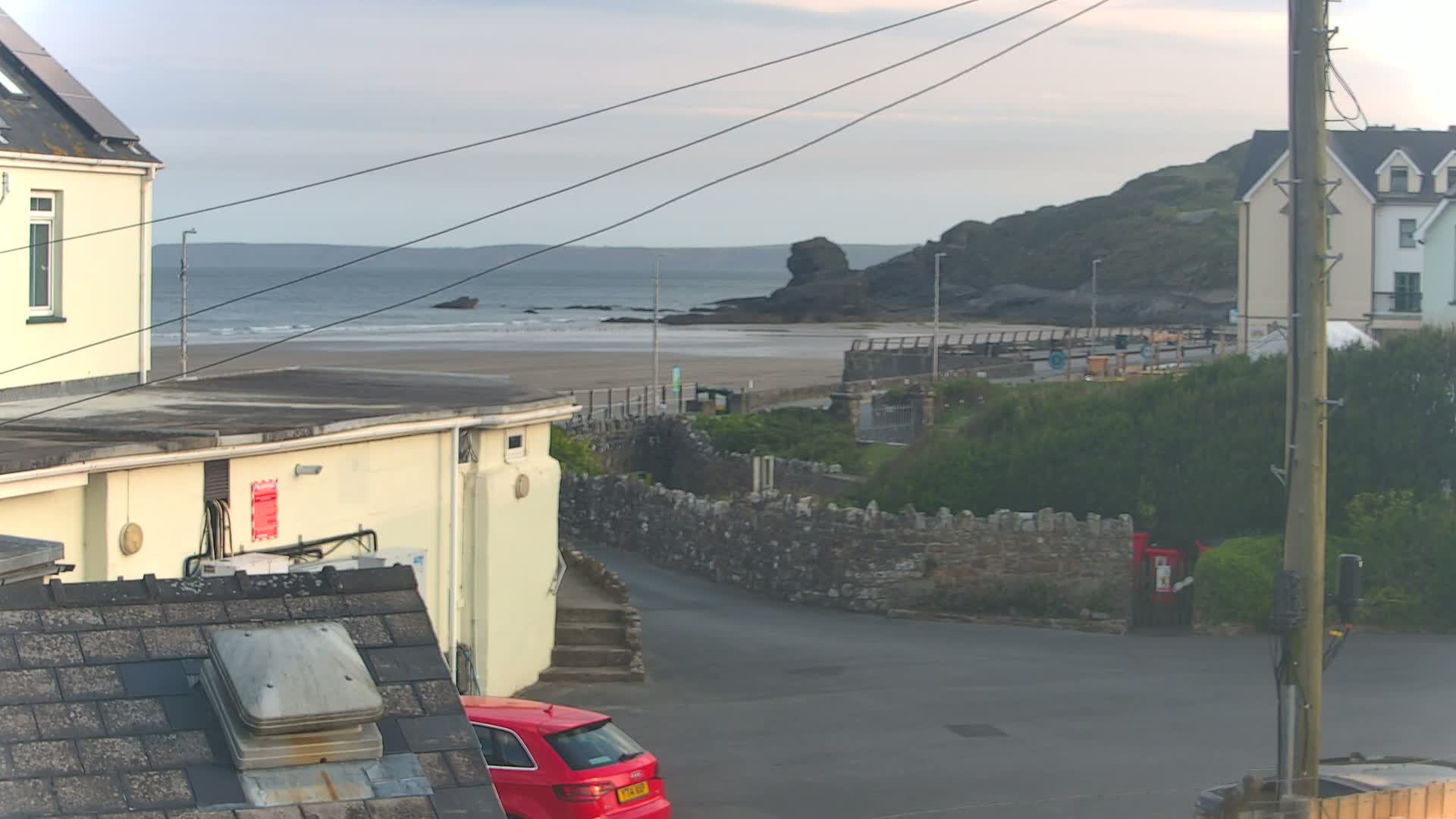 A view from a rooftop shows a sandy beach with dark rocks and calm ocean under an overcast sky, with buildings and a red car in the foreground.