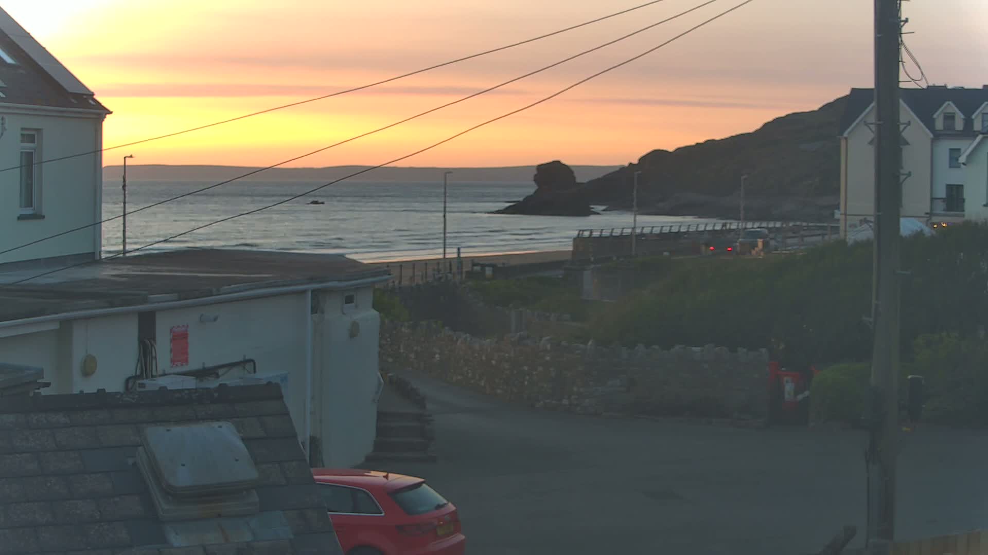 A sunset over a calm ocean, viewed from between houses, with a red car parked in the foreground.