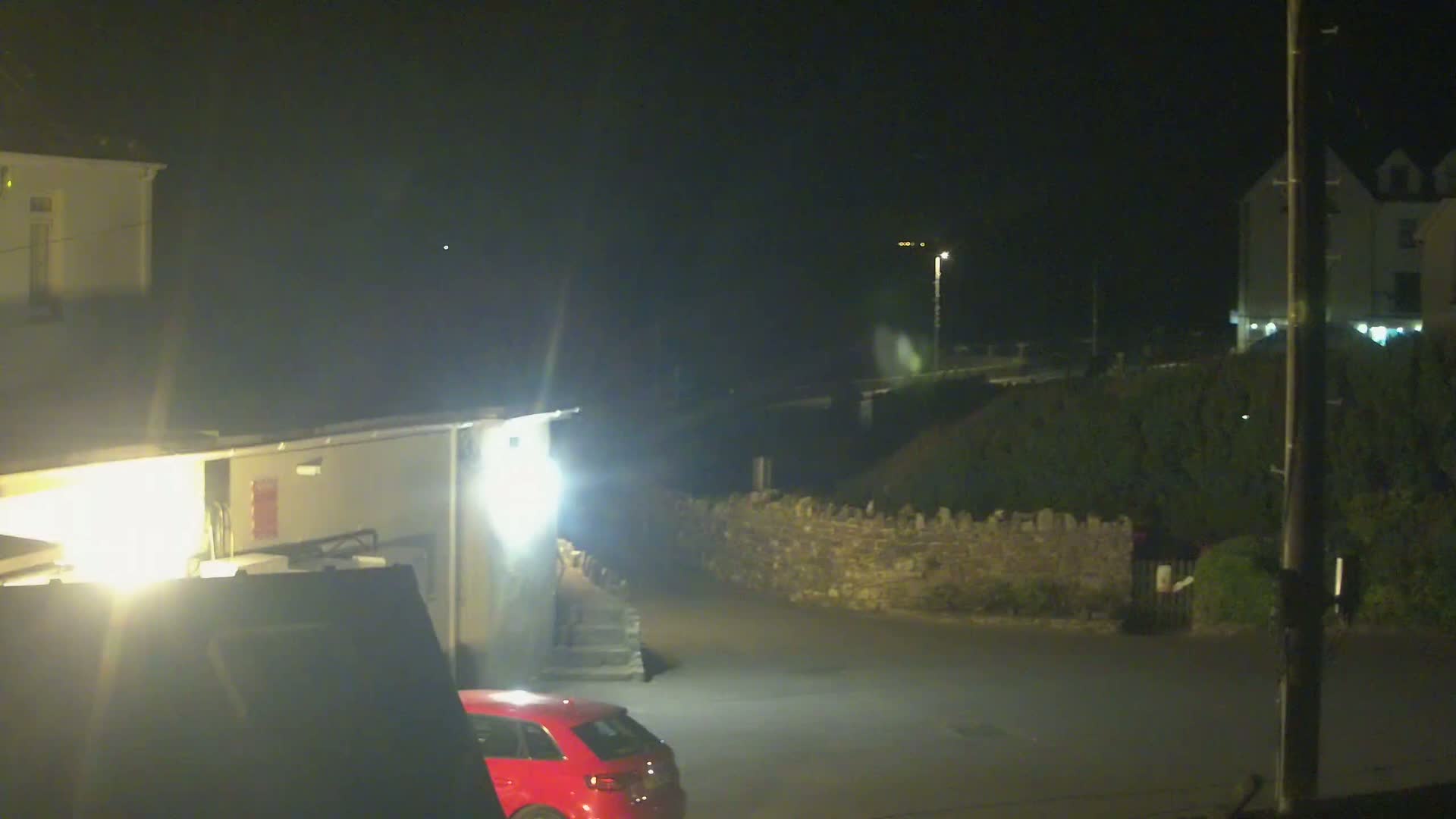 A nighttime view shows a red car parked near a building, a low stone wall, and other buildings in the distance under a dark sky.