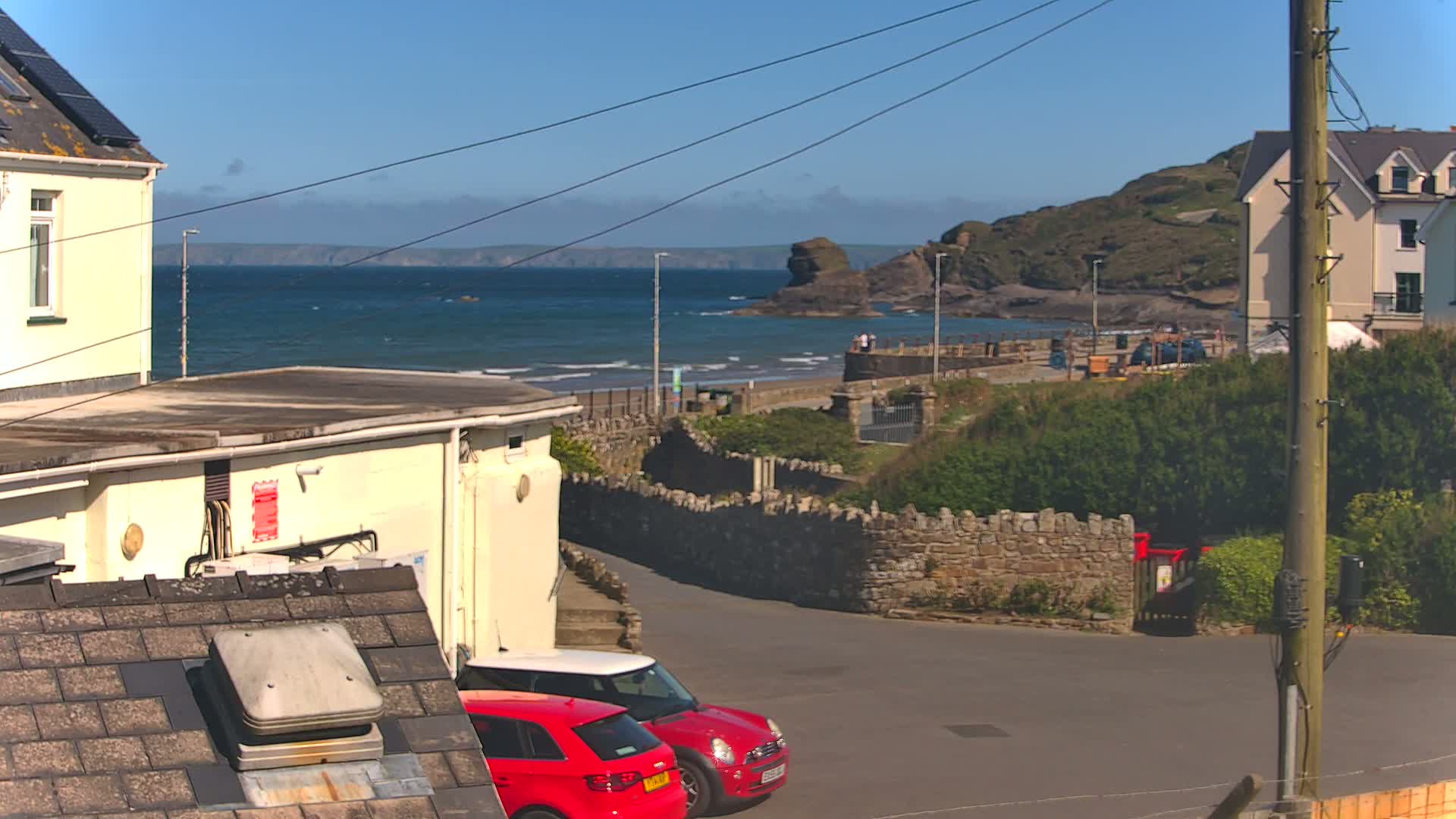 A sunny coastal scene shows a section of a sandy beach with small waves, a rocky headland, a stone wall, parked cars, and buildings.