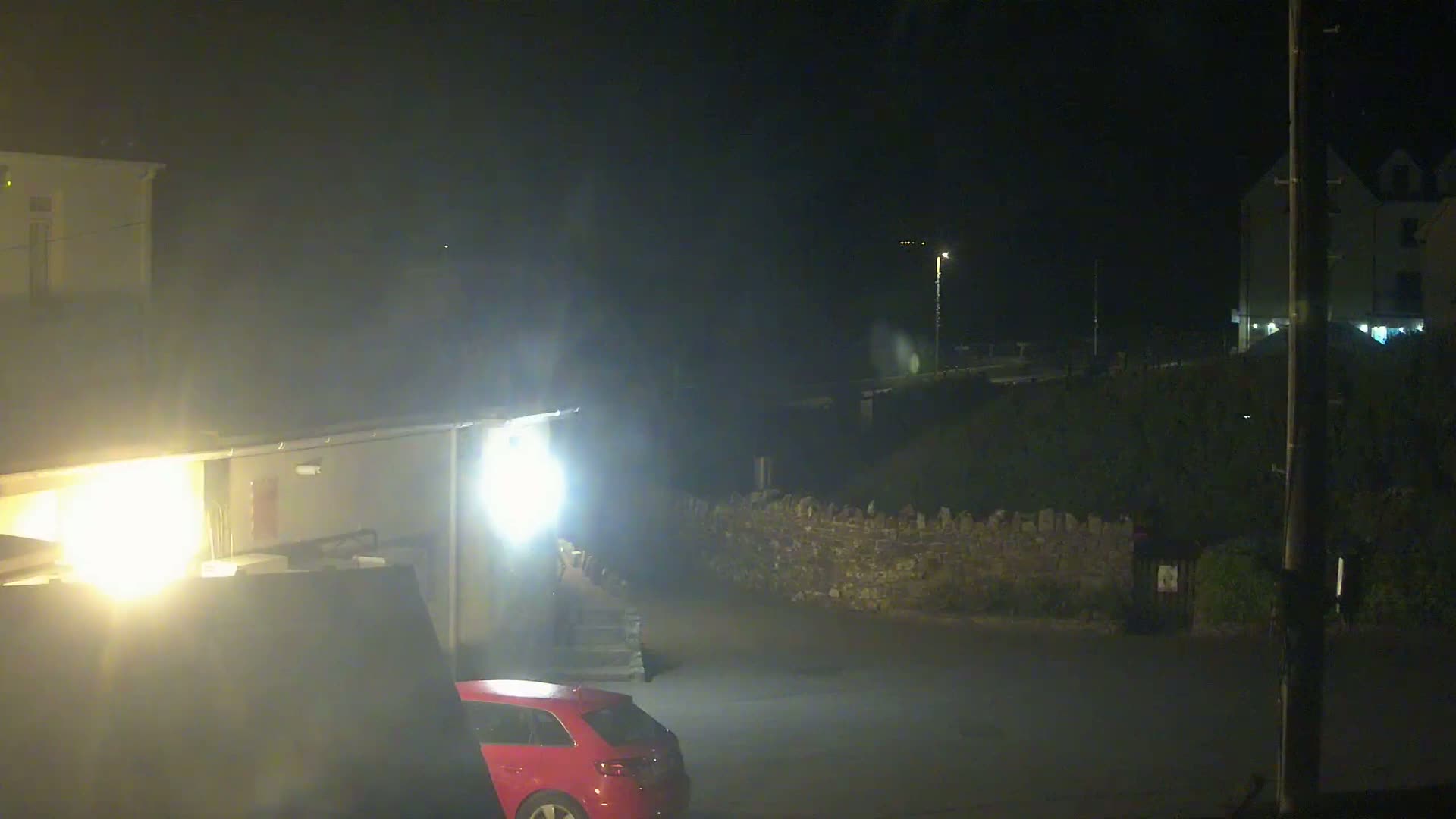 A red car is parked in a dimly lit area at night near buildings and a low stone wall.