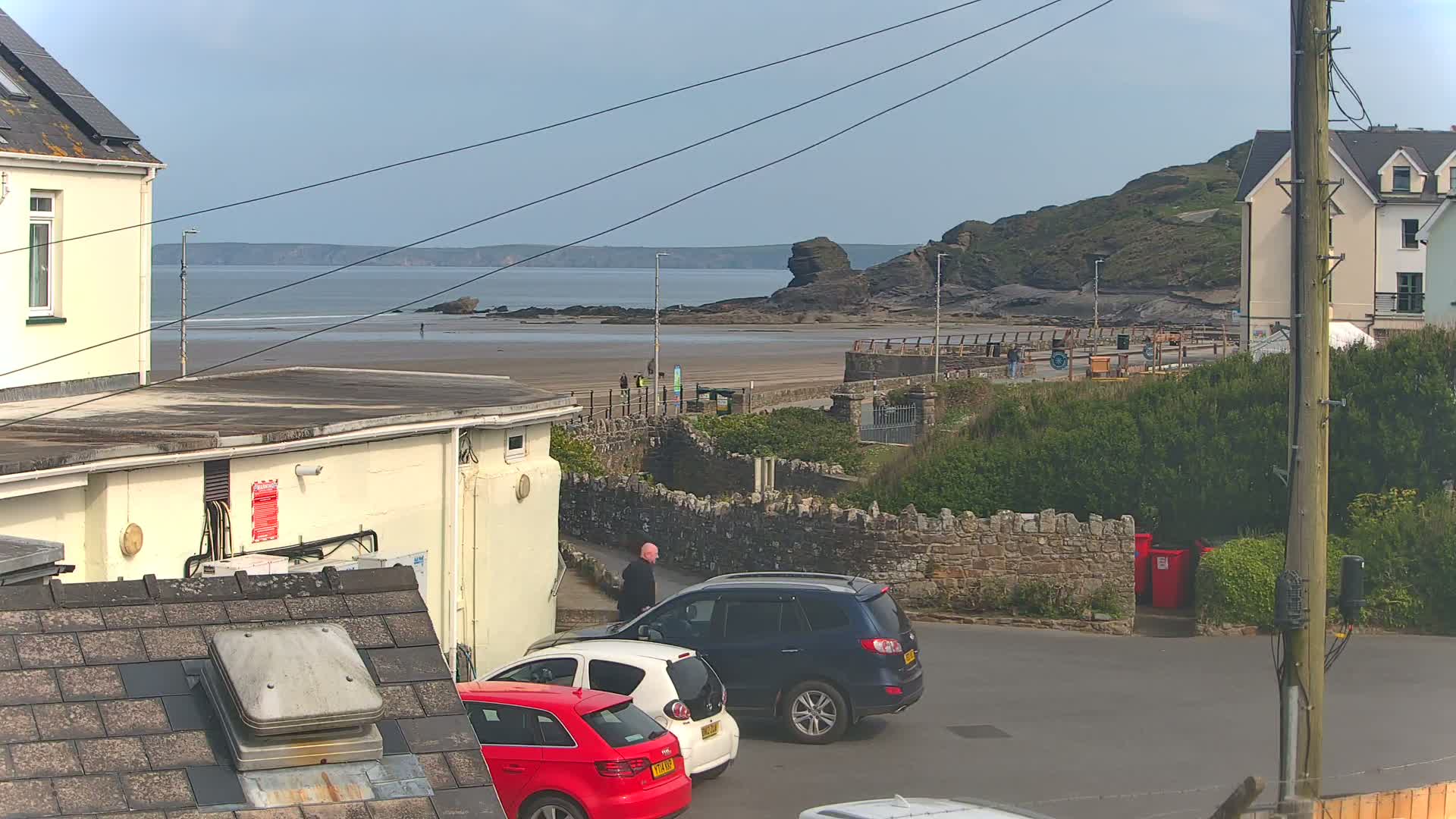 A seaside view shows a sandy beach with a few people on it, a rocky outcrop in the water, buildings, and parked cars, all under a mostly clear sky.