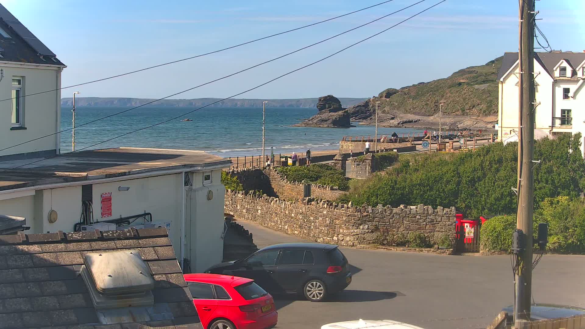 A seaside town scene on a sunny day shows a calm ocean, sandy beach with a few people, and parked cars near stone walls and buildings.