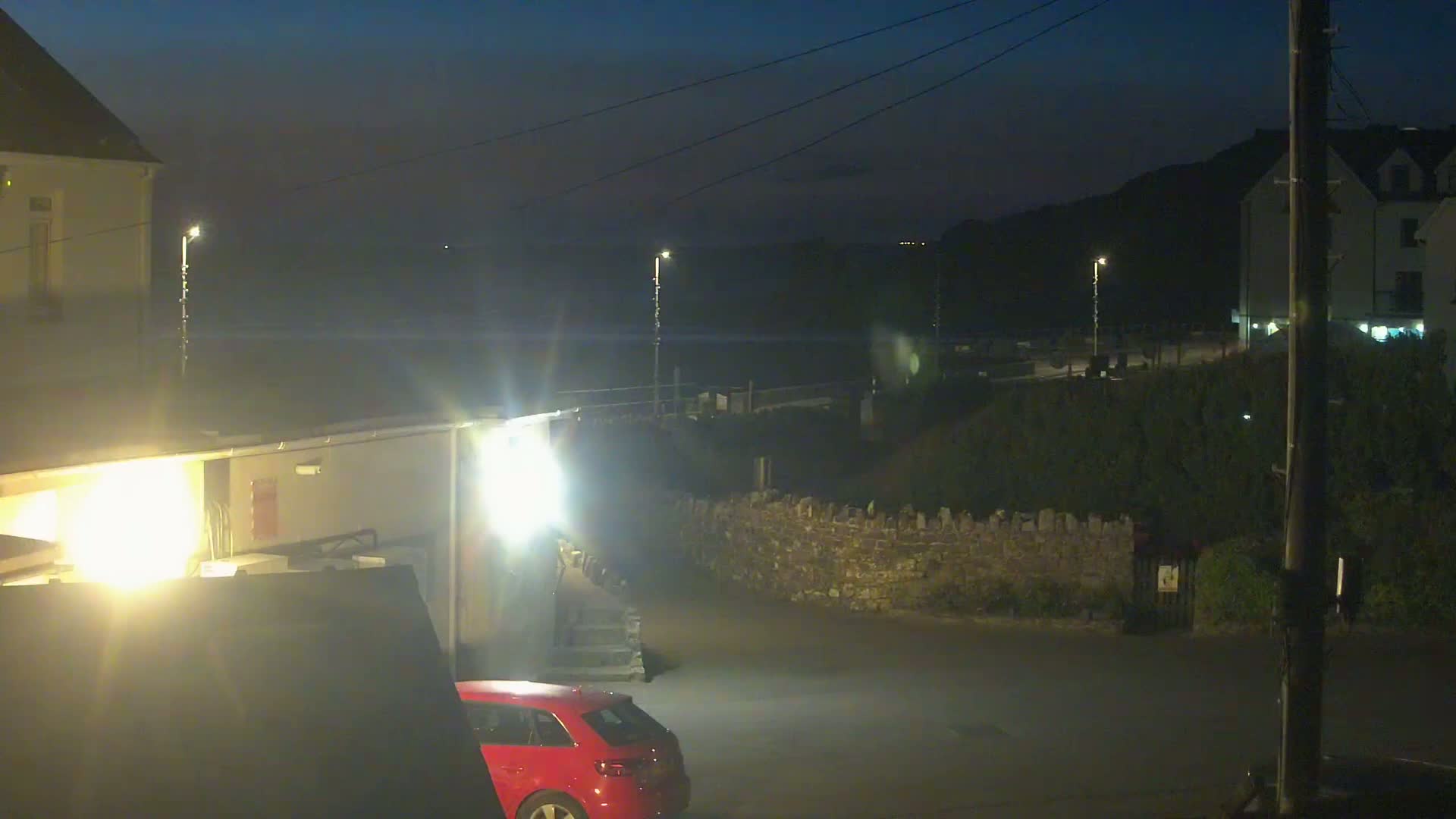 A red car is parked near a building at night, with a low stone wall and dark, possibly coastal, scenery in the background under a dark, clear sky.
