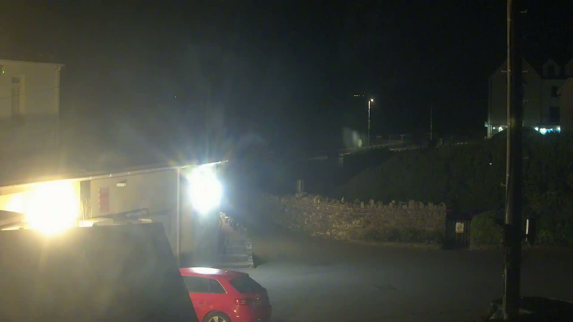 A red car is parked in a dimly lit area at night near a low stone wall and buildings, under hazy or foggy conditions.