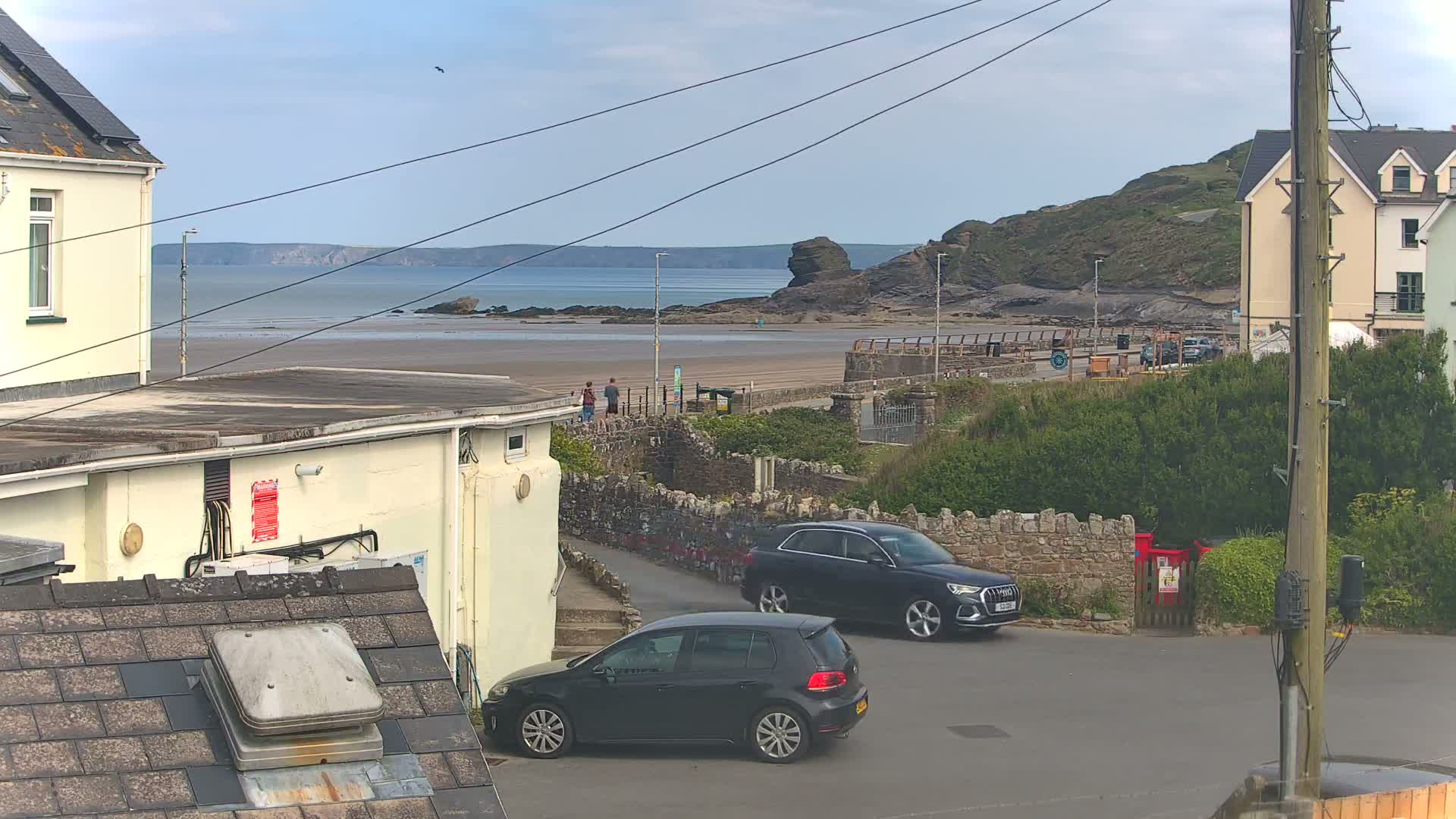 Overlooking a sandy beach and calm ocean from a residential area, two dark-colored cars are parked near a stone wall under a mostly clear sky.