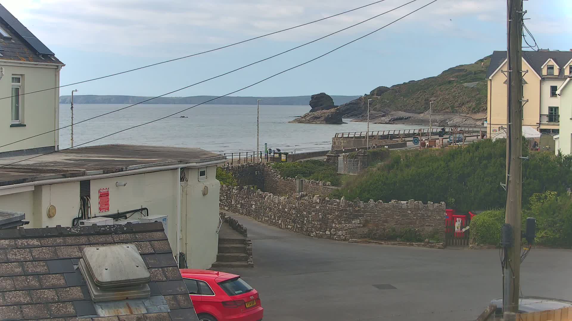 A seaside view shows a calm ocean under an overcast sky, a rocky coastline with a few people on the beach, buildings, and a red car parked near a stone wall.