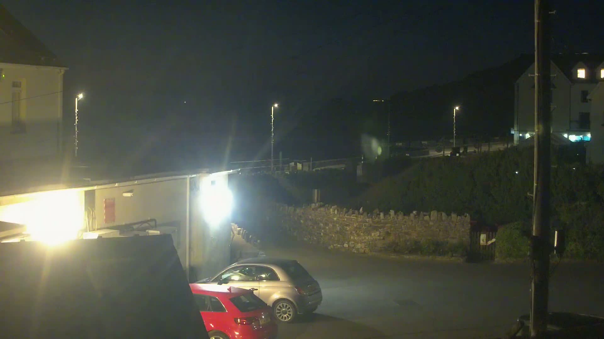 At night, two cars are parked in a dimly lit area next to a low stone wall, with buildings and streetlights visible in the background.