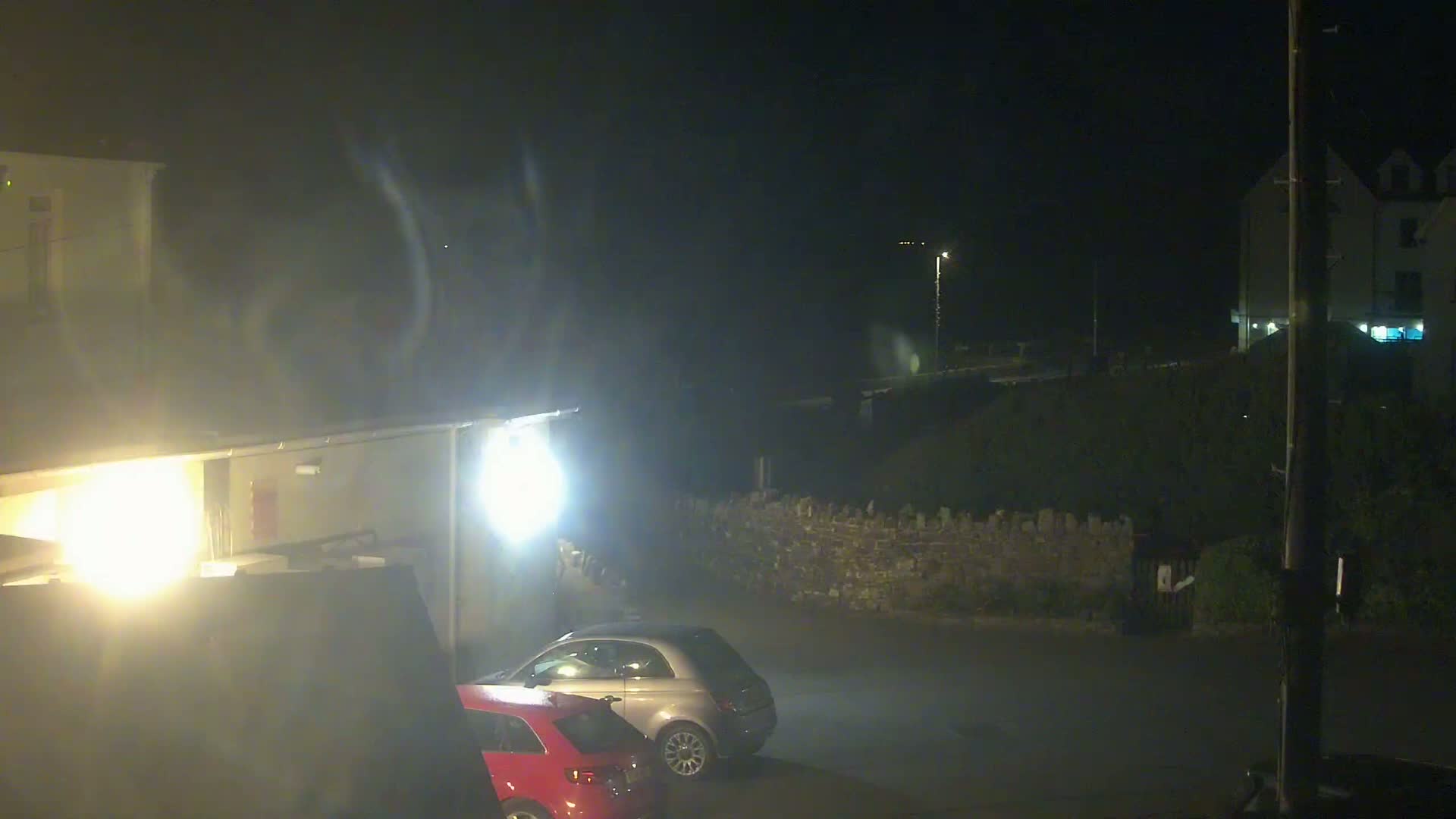 At night, under hazy conditions, two cars are parked in a lot next to a low stone wall and buildings.