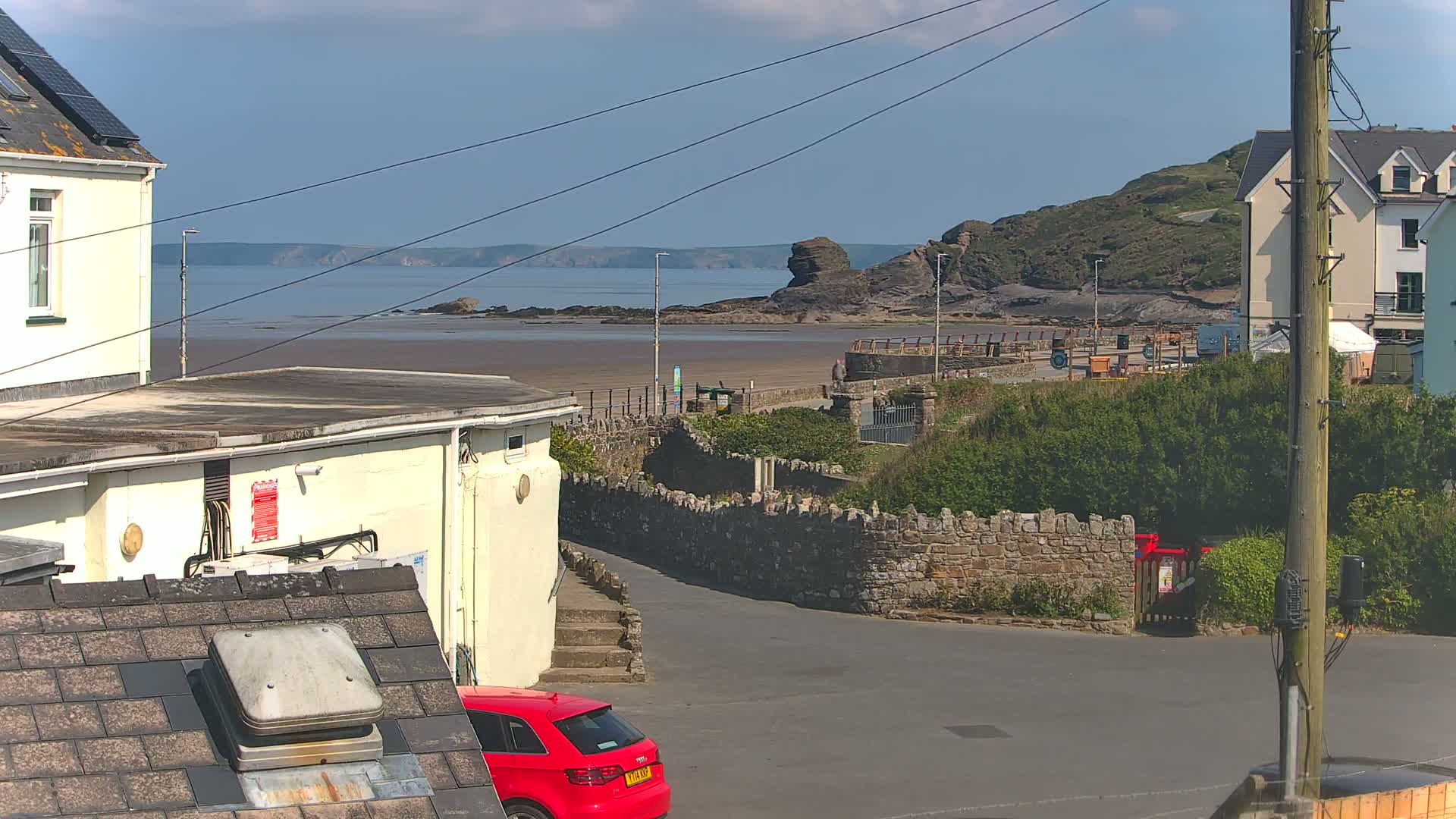 A sunny view of a coastal town shows a sandy beach, a rocky outcrop in the sea, buildings, and a red car parked near a stone wall.