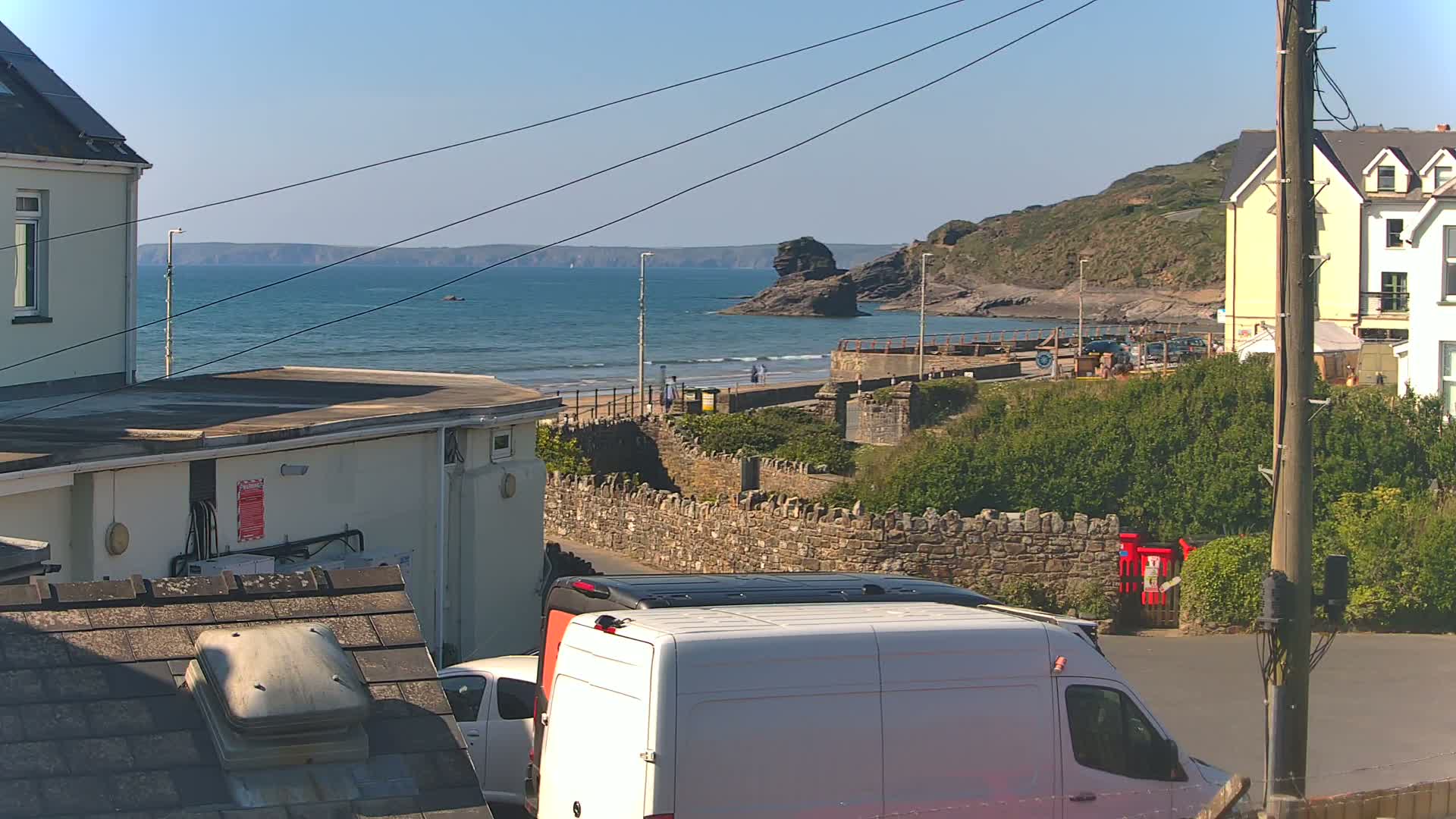 A sunny seaside view shows a calm ocean with a rocky outcropping, a sandy beach with a few people walking, buildings on a hillside, and a white van parked on a street.