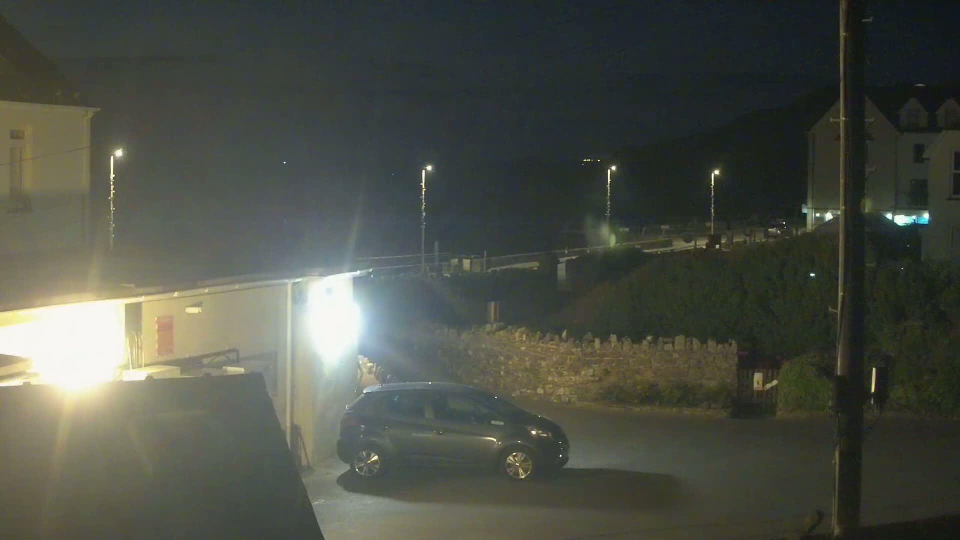 A dark-colored car is parked in a nighttime lot next to a low stone wall, with buildings and lights visible in the background under a dark sky.