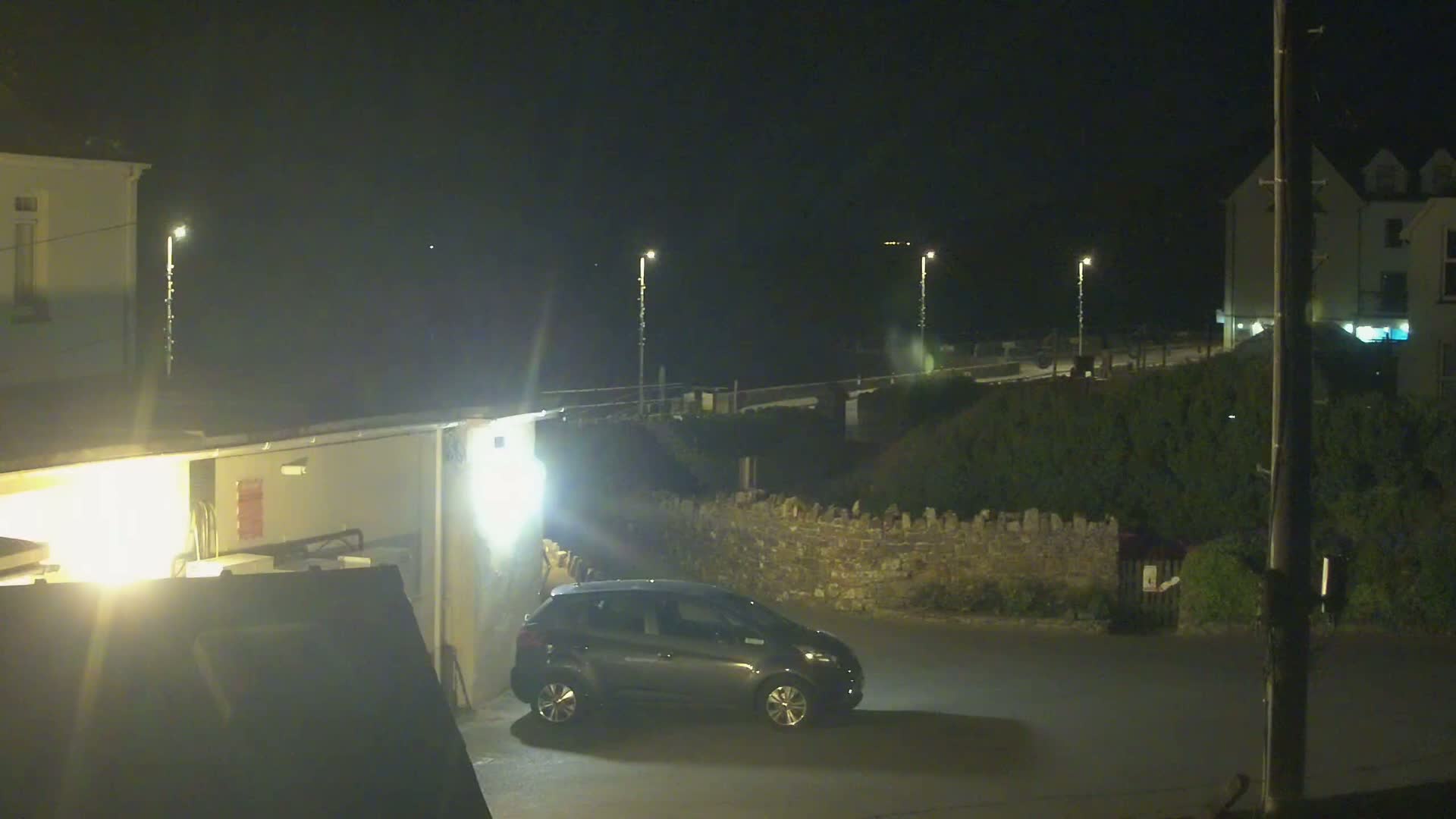 A dark gray car is parked in a dimly lit area at night, next to a low stone wall and buildings.